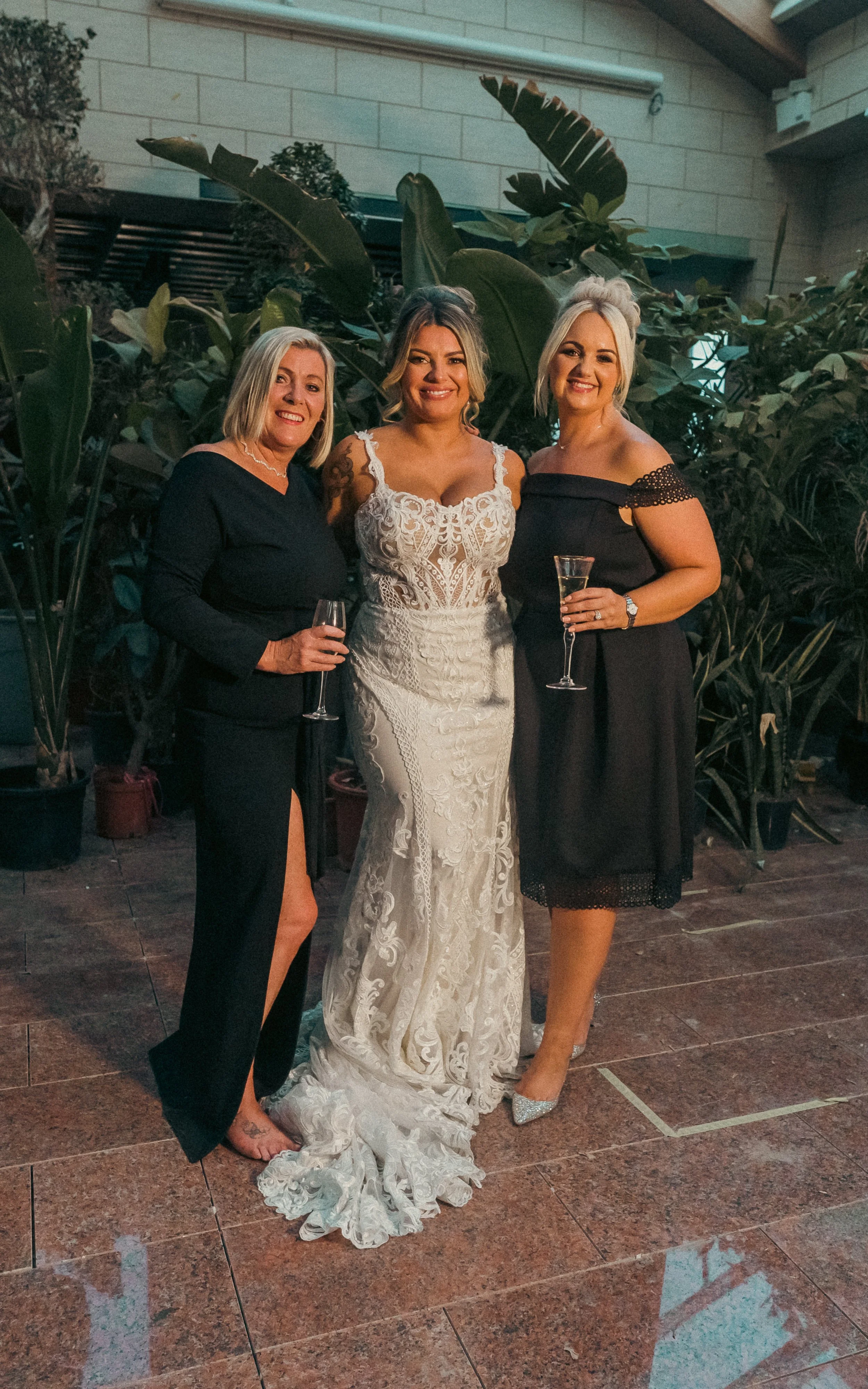 Three women dressed elegantly standing together indoors, celebrating with drinks, with large plants and a stone wall background.