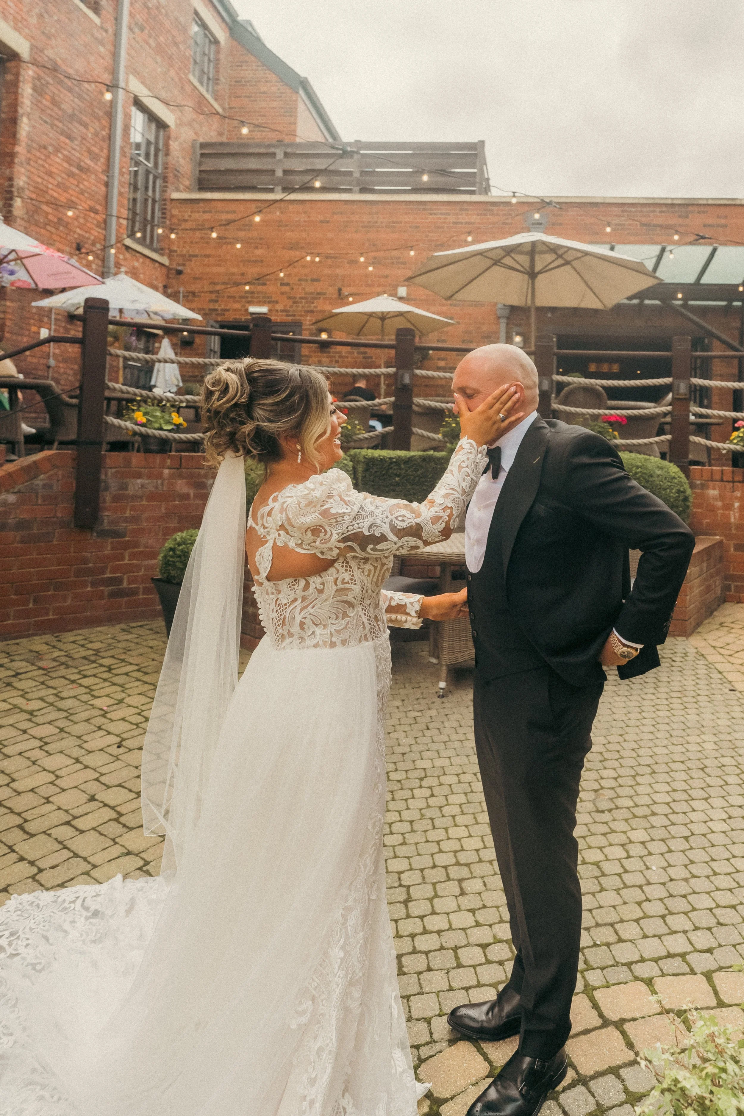 Bride in a lace wedding dress touching groom's face, smiling, on a brick patio with string lights and umbrellas.