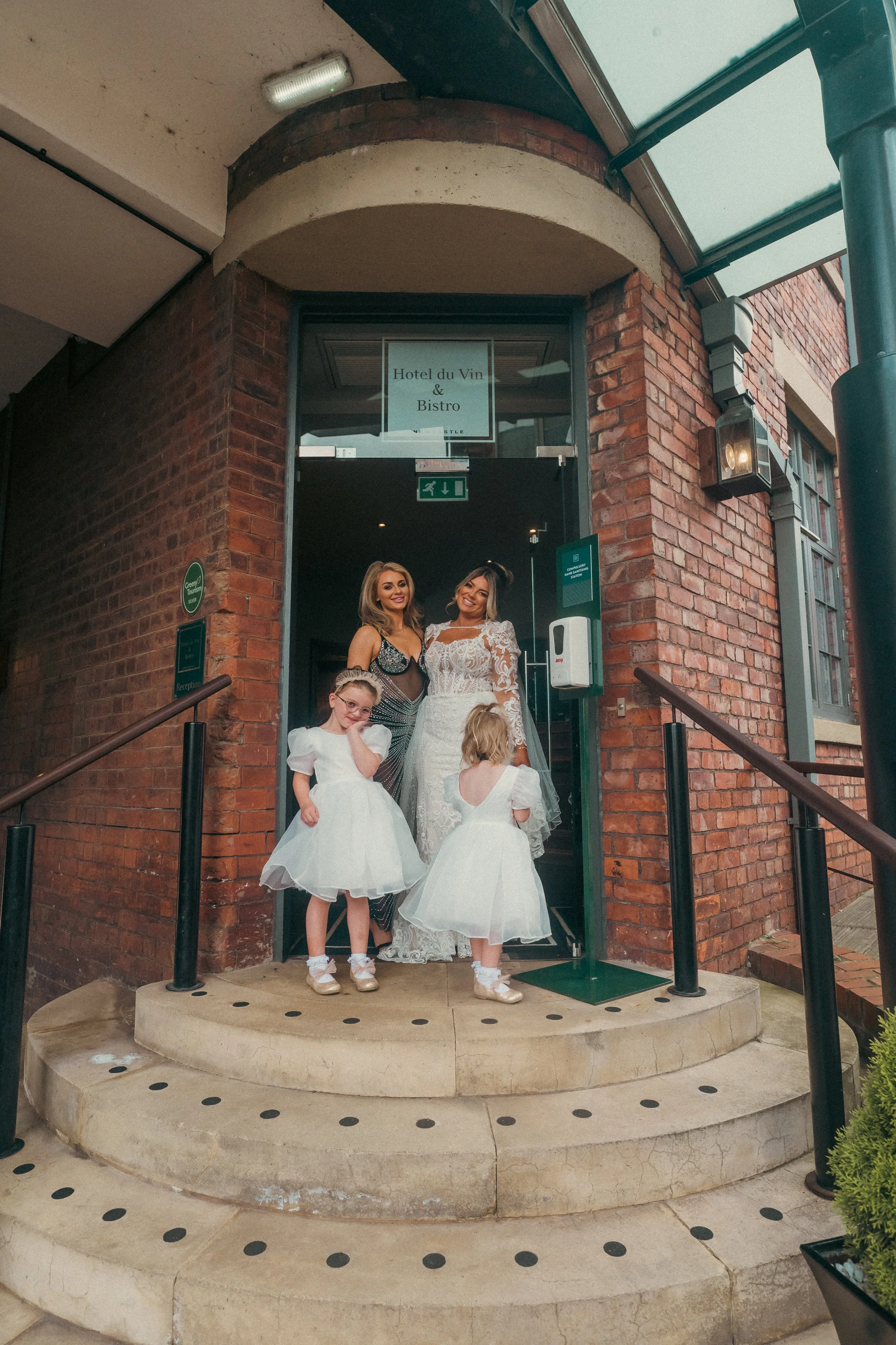 Two women and two young girls standing at the entrance of a brick building, with a sign reading 'Hotel du Vin & Bistro' above them. The women are in elegant dresses, and the girls are in white dresses, possibly for a special occasion or wedding.