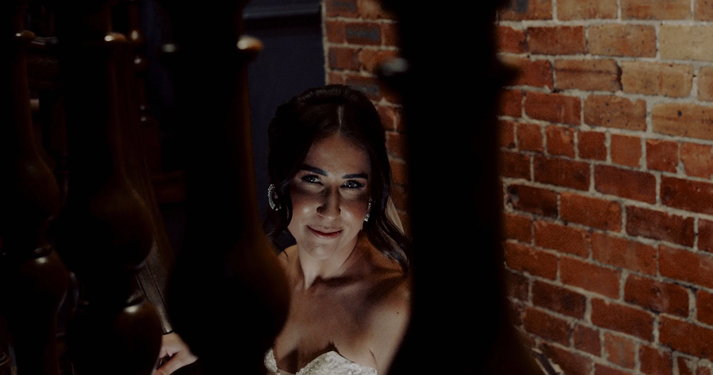 A woman smiling in a dimly lit room with a brick wall background, seen through the gaps in a wooden staircase railing.