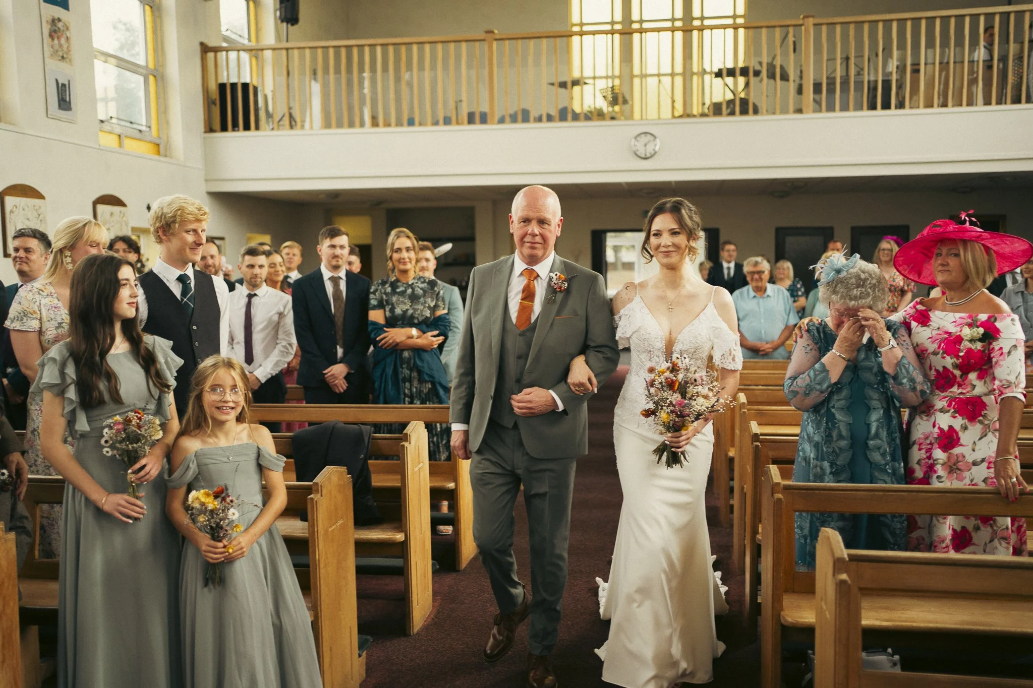 A bride wearing a white wedding dress walking down the aisle with an older man, possibly her father, at a wedding ceremony inside a church filled with seated and standing guests. Some guests are dressed in formal and floral attire, with one woman wea