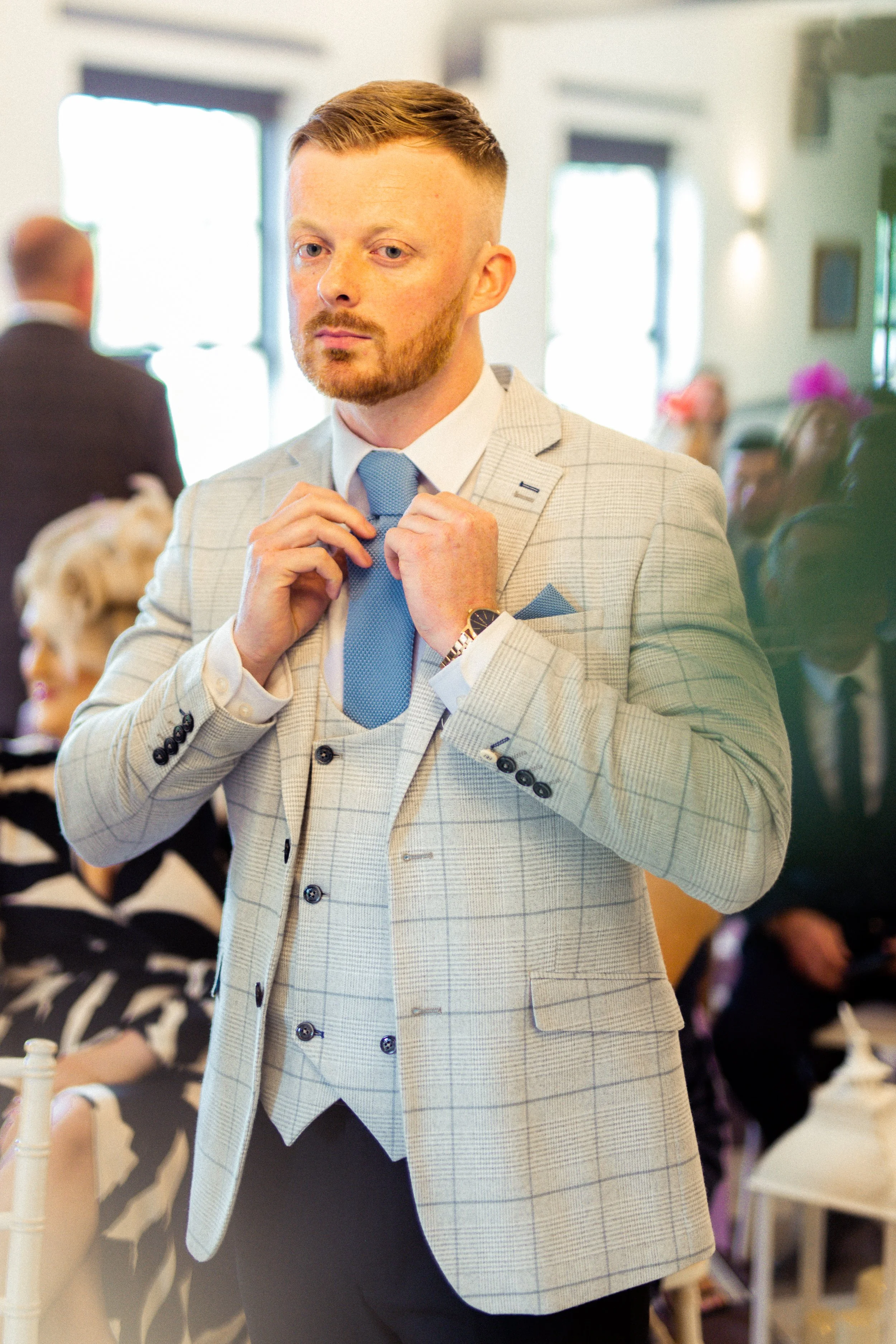 A young man adjusting his light blue tie at a formal indoor event, dressed in a beige checkered suit vest and blazer, with other guests visible in the background.
