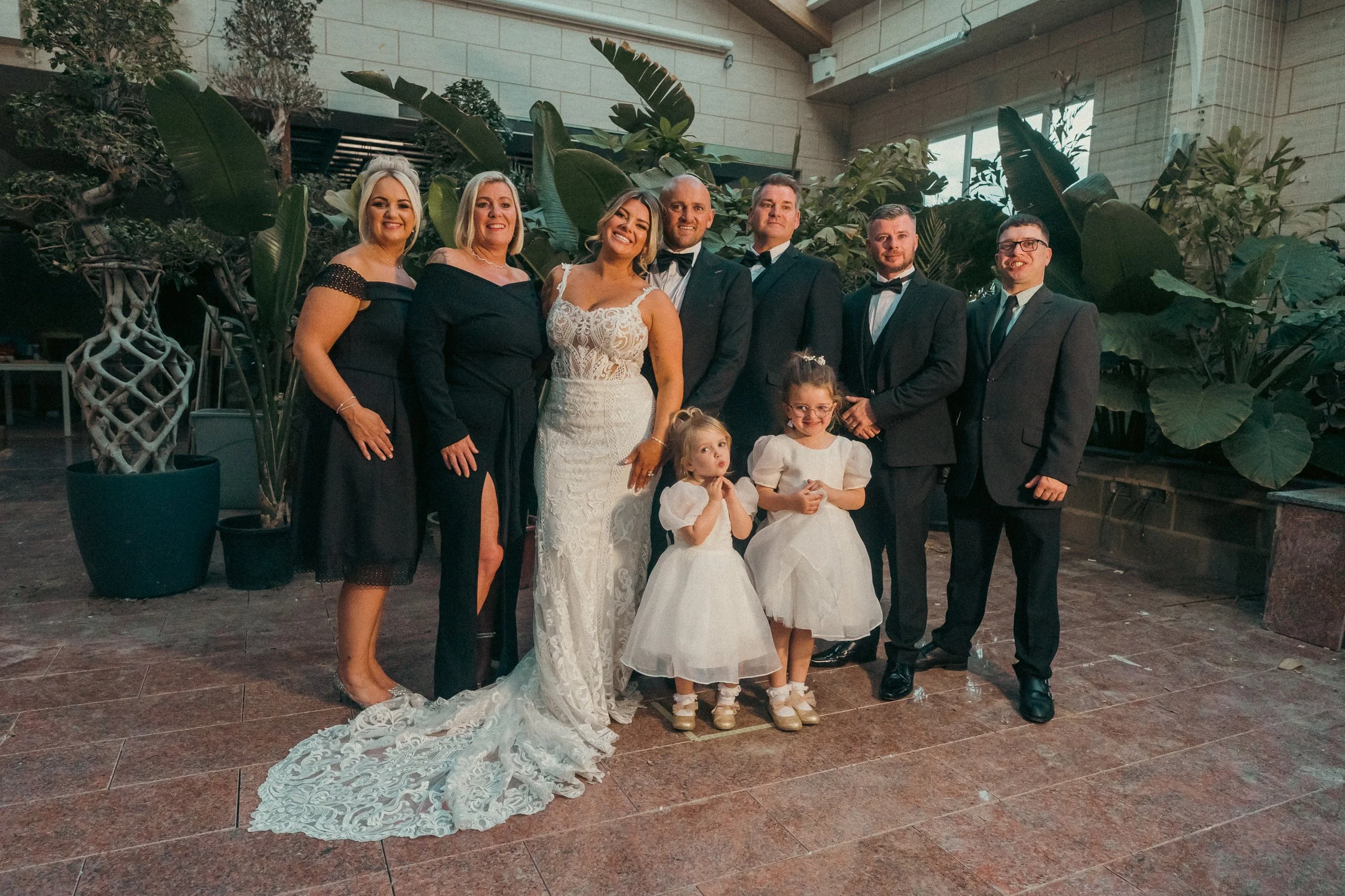 A group of adults and children dressed in formal attire standing together indoors, with large green plants in the background, likely at a wedding celebration.
