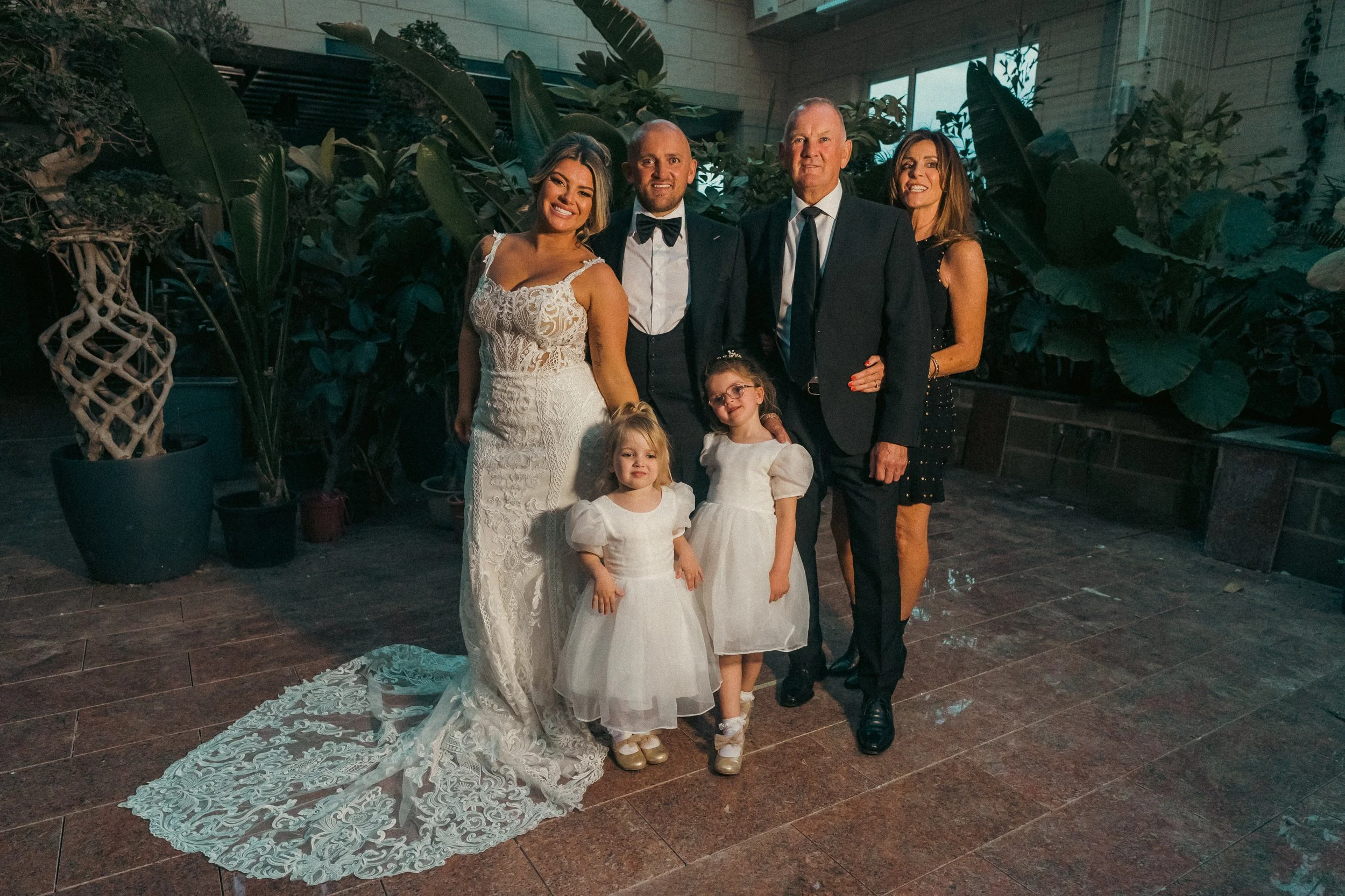 A group of six people, including a bride in a white lace wedding dress, a groom in a tuxedo, and four children, standing together in an indoor setting with lush green plants in the background.
