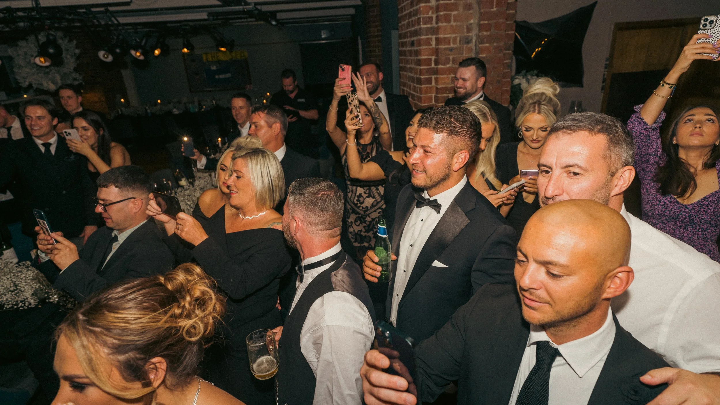 People in formal attire at a lively party or celebration, some taking photos or videos with smartphones, with warm lighting and brick walls in the background.