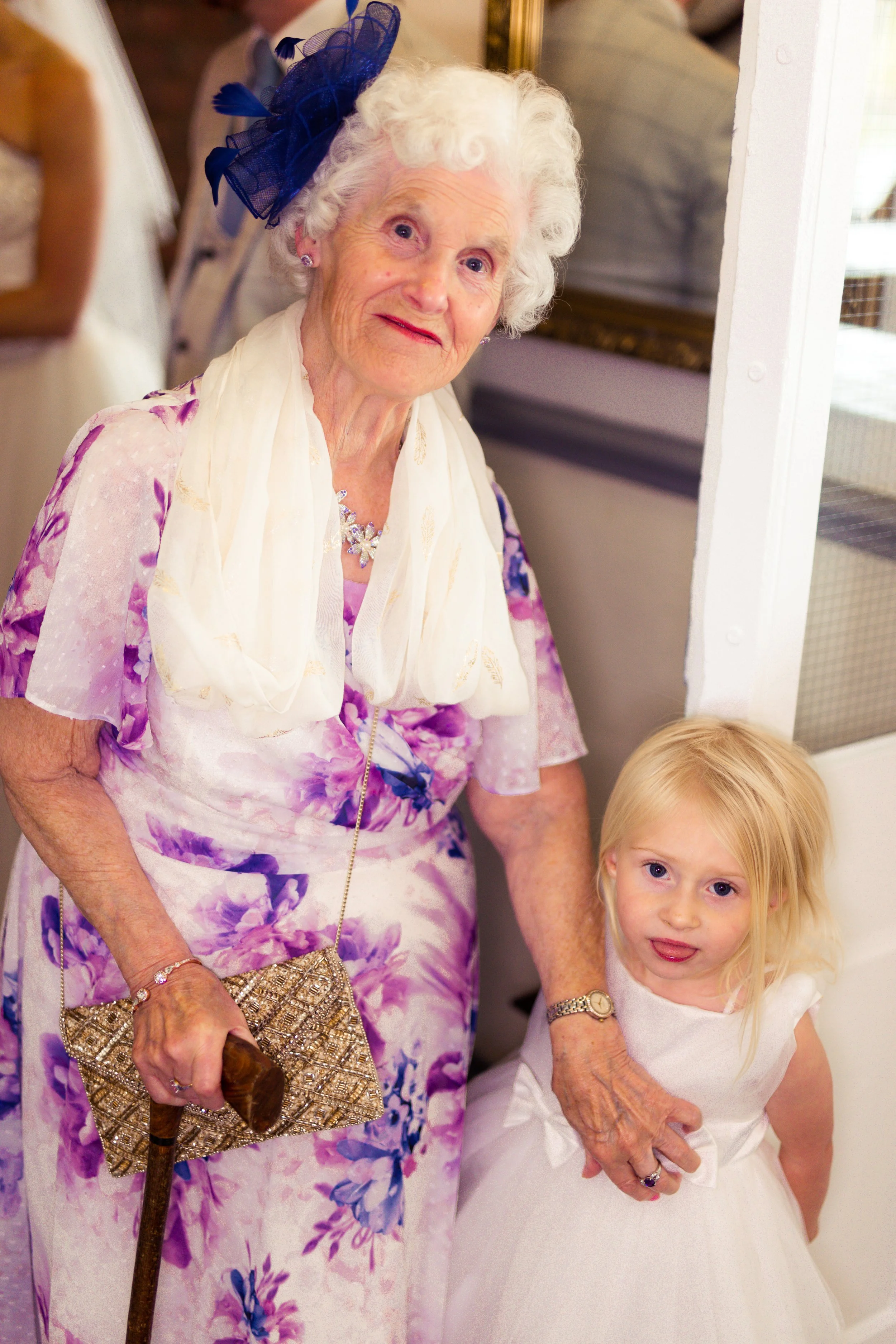 An elderly woman with white curly hair wearing a purple floral dress, a cream scarf, and a dark blue fascinator, standing indoors next to a young girl in a white dress. The elderly woman is holding a decorative clutch and a walking cane, and the girl