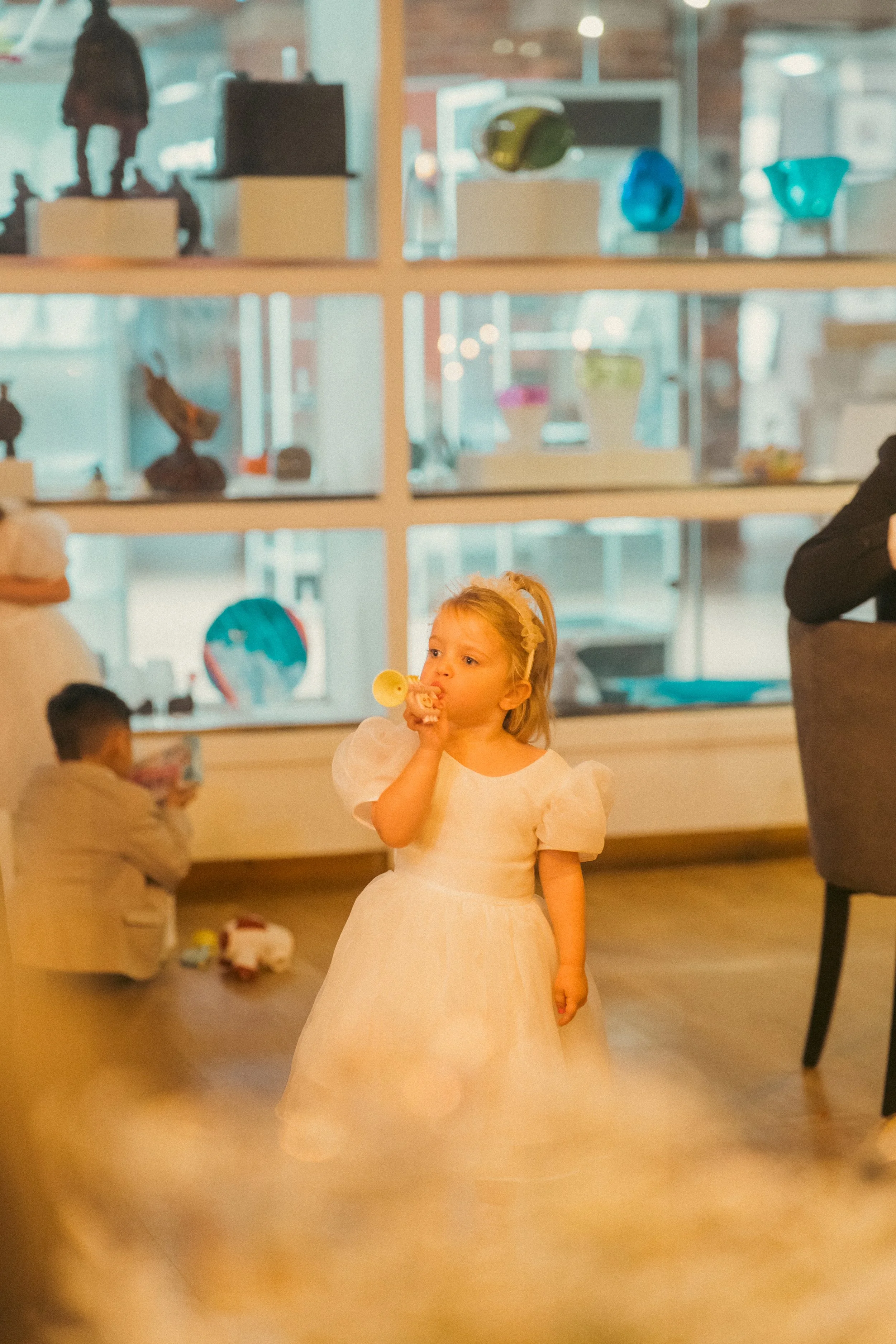 Young girl in a white dress eating a snack in a room with large display shelves filled with colorful art glass sculptures.