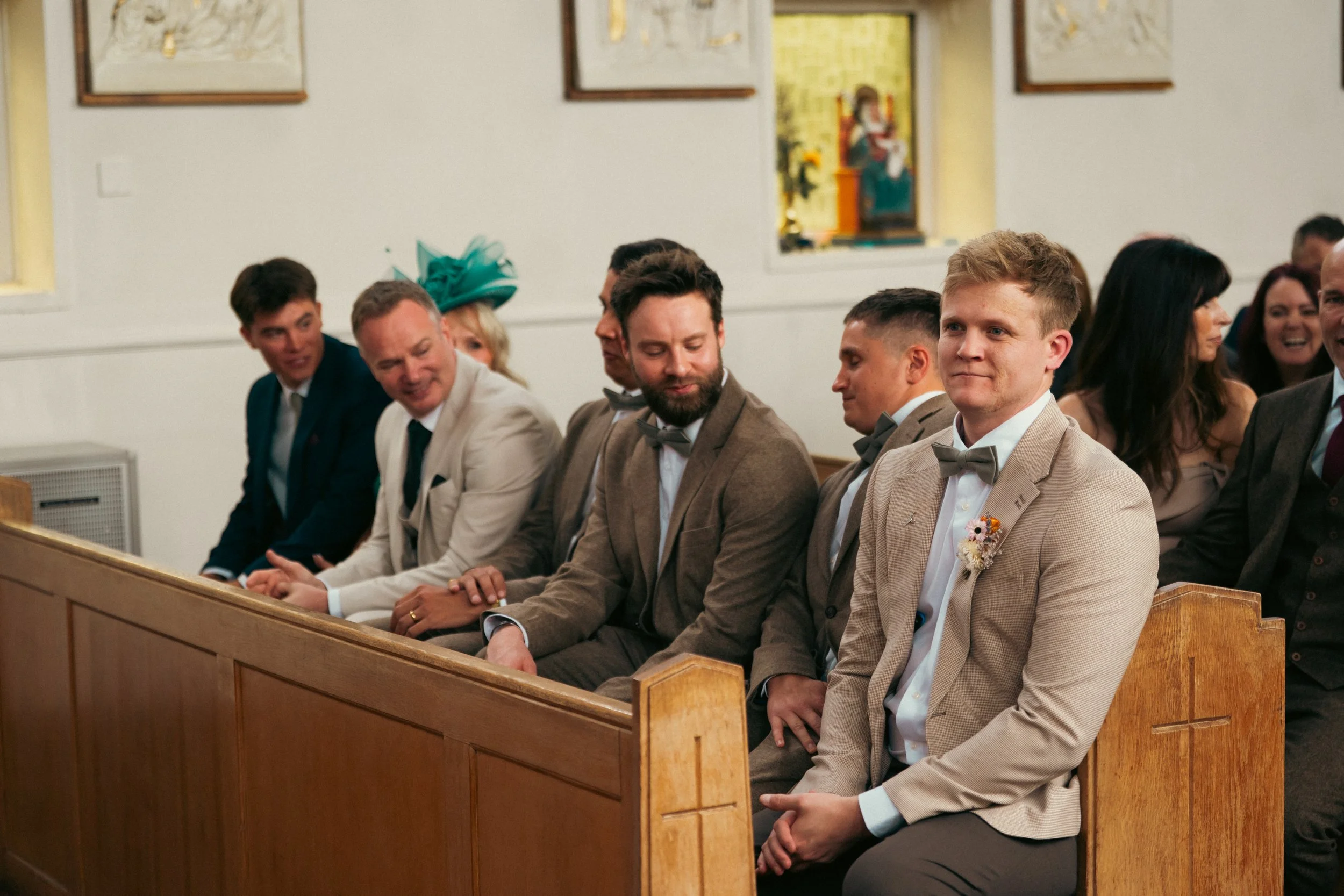 Group of people sitting in church pews during a wedding ceremony, dressed in formal attire, some smiling and talking.