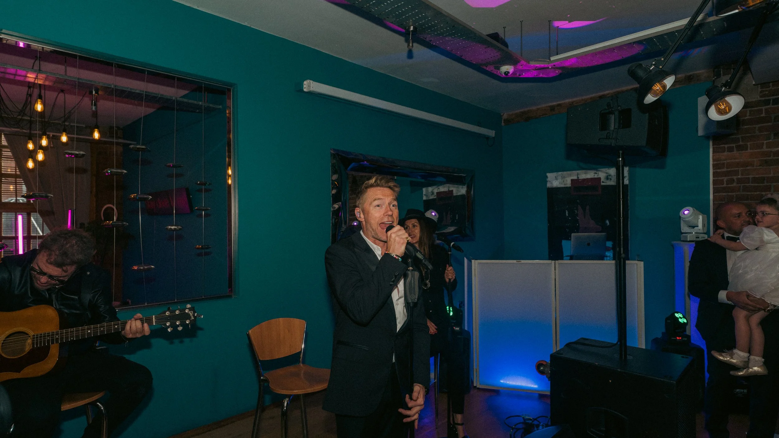 A man in a black suit singing into a microphone on a stage in a dimly lit room, with a guitarist to his left and a woman in the background holding a microphone. To the right, a man in a suit is holding a young girl dressed in white, with colorful sta