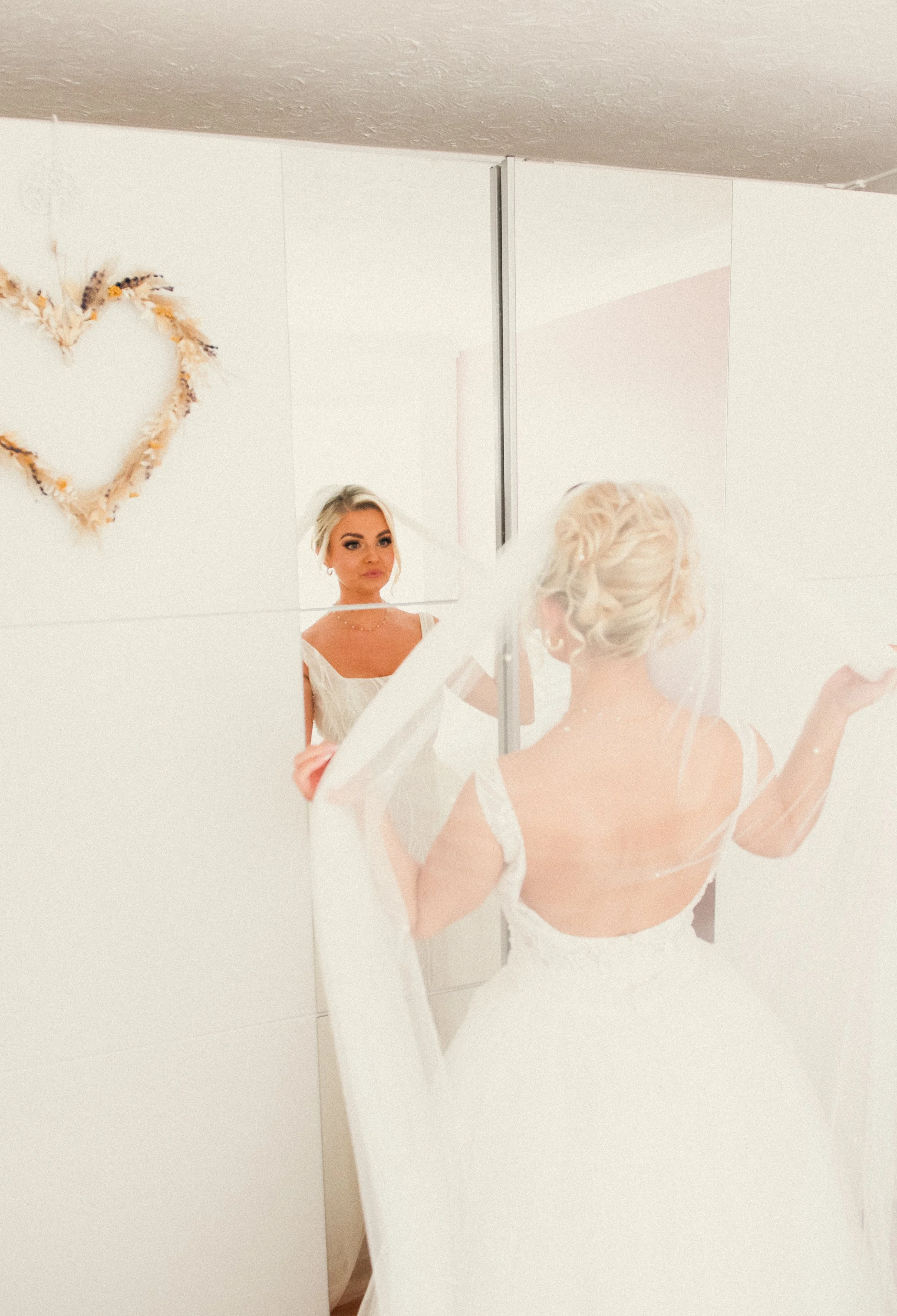 A bride in a white wedding dress looks at her reflection in a mirror, with a heart-shaped decoration on the wall behind her.