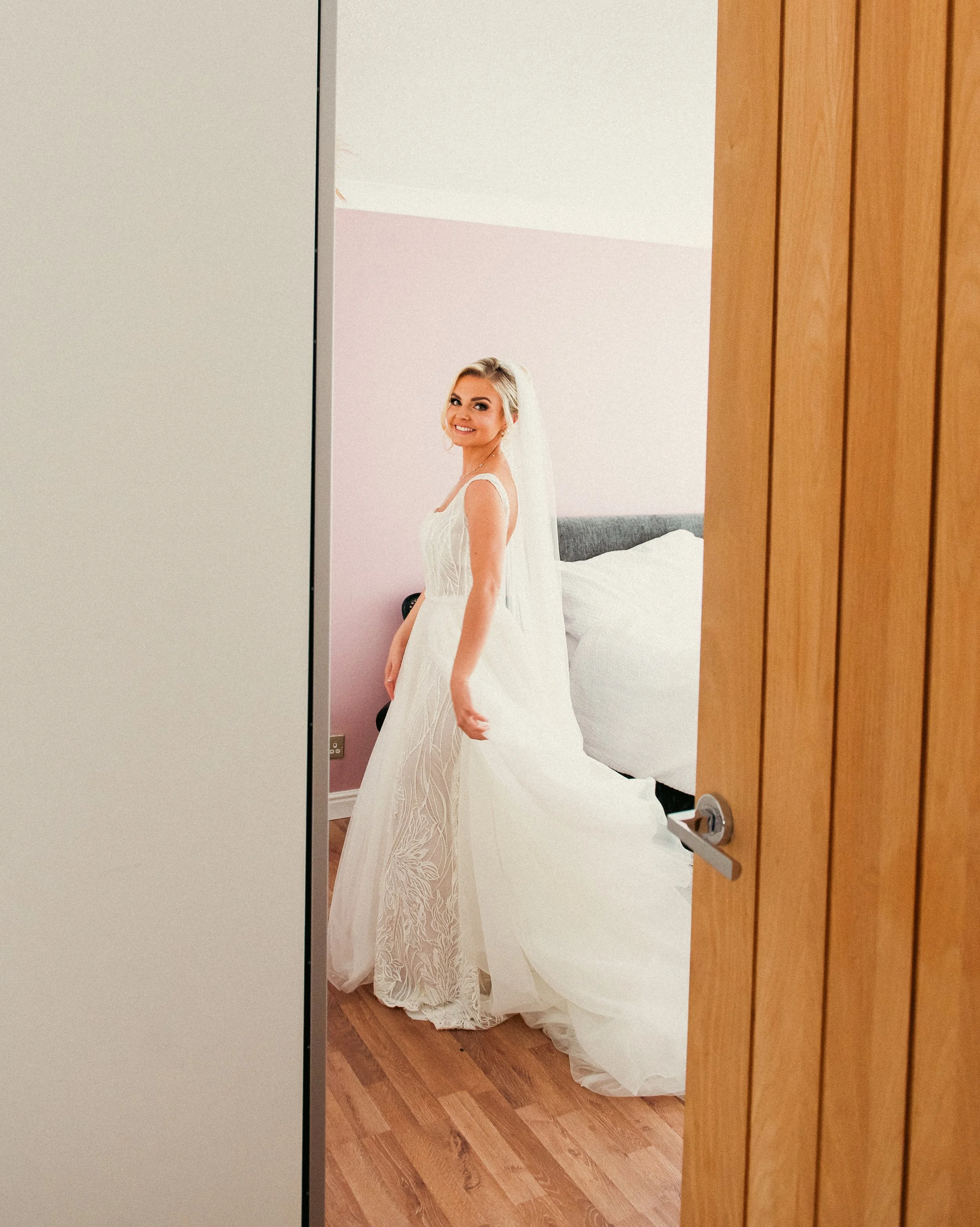 A bride in a white wedding dress with lace details, smiling and looking at the camera, standing in a bedroom seen through an open door.