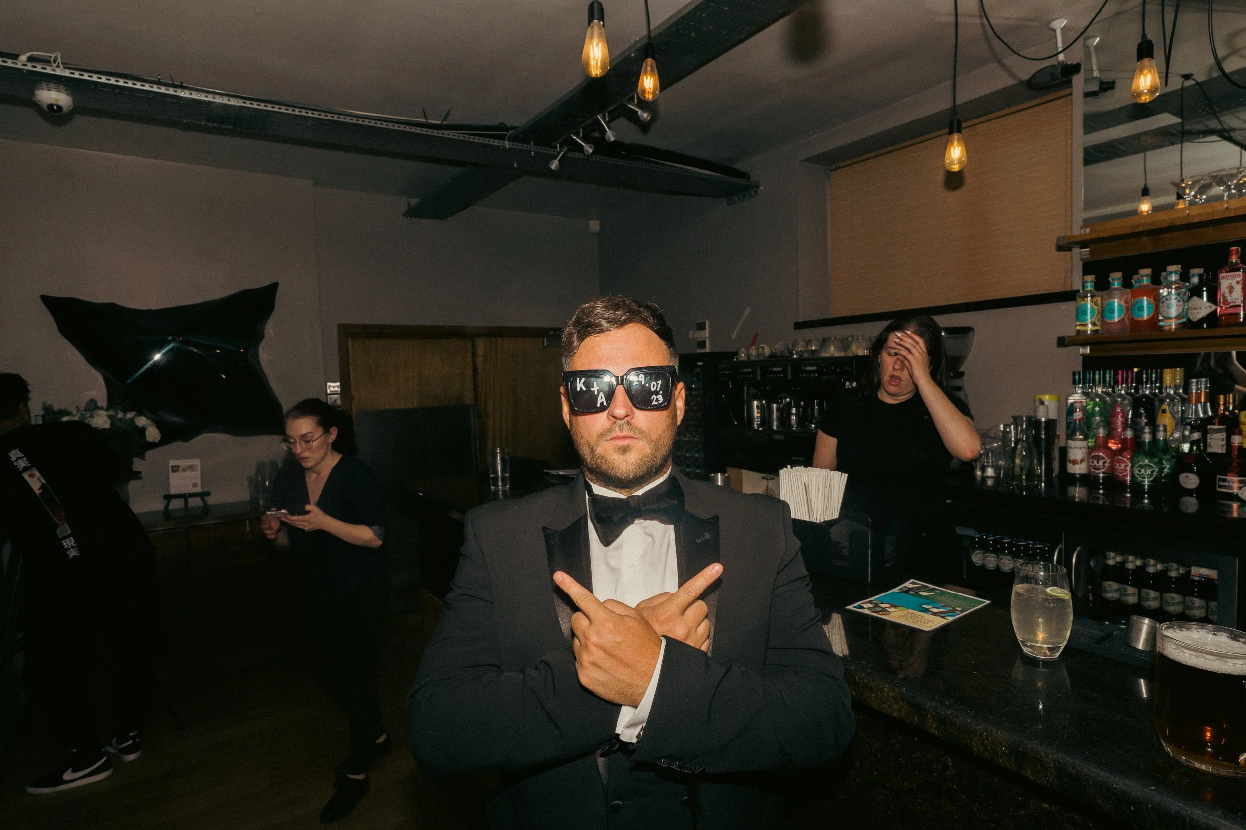 Man in tuxedo and sunglasses crossing arms in a bar with two women and bar equipment in the background.