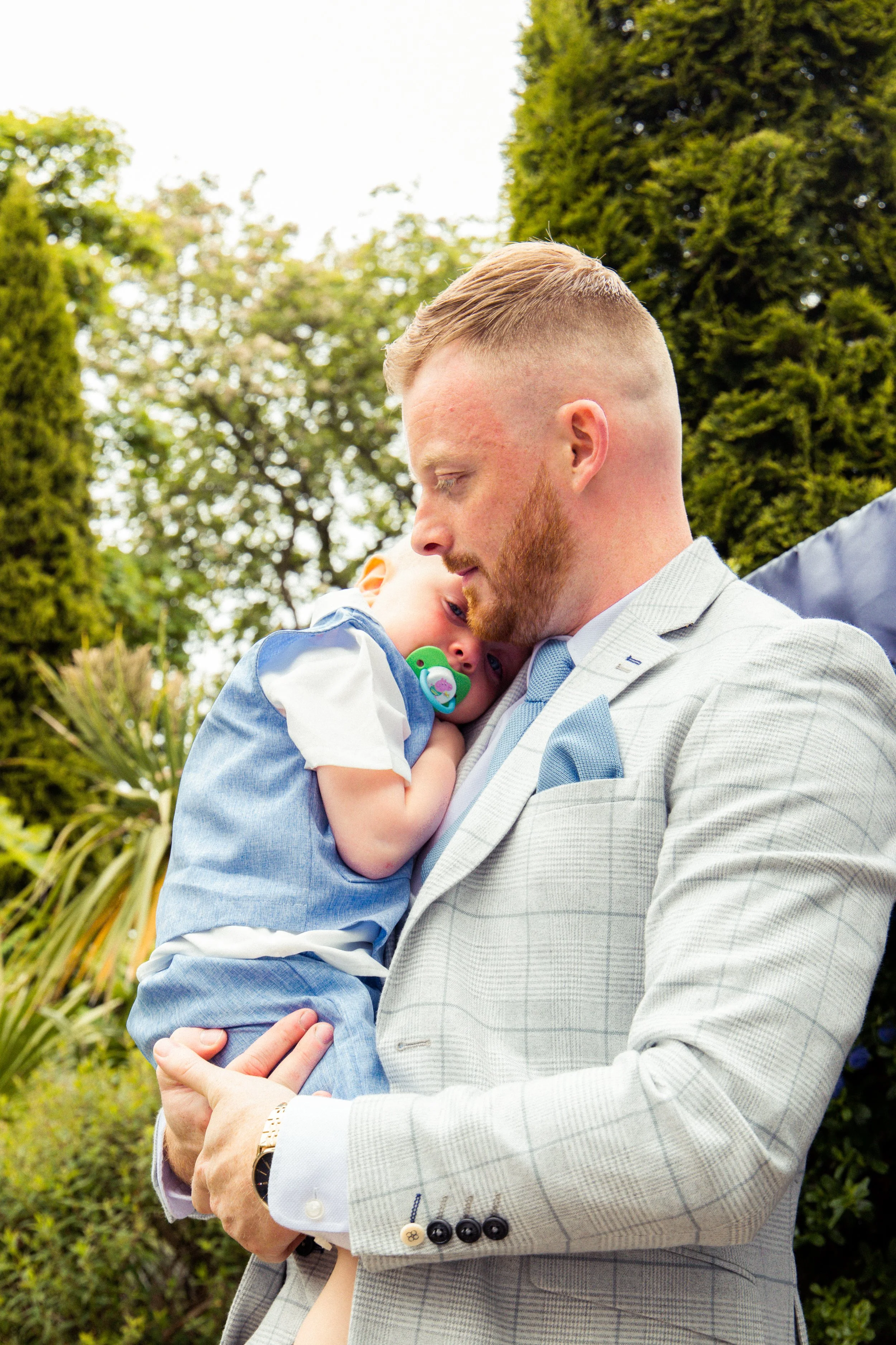 A man in a light gray plaid suit holding a young boy with a pacifier outdoors among trees and greenery.