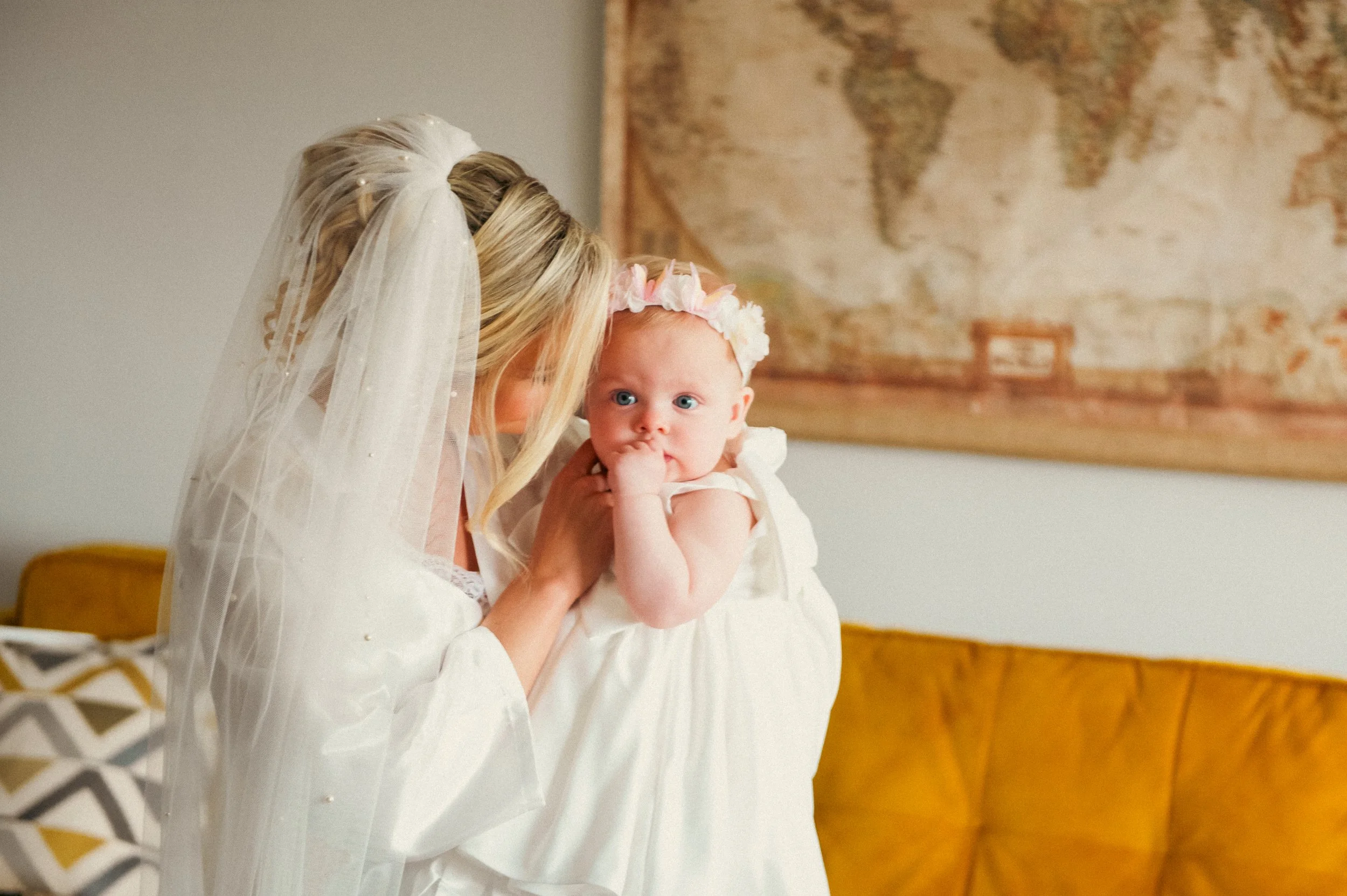 A bride wearing a wedding dress and veil holding and comforting a baby girl dressed in white with a floral headband, indoors with a world map on the wall and a mustard-colored sofa