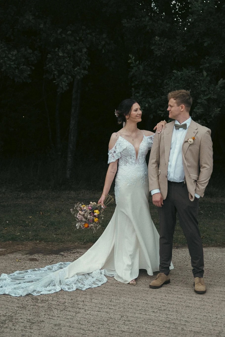 A bride and groom stand together outdoors at sunset, looking at each other. The bride wears a white lace wedding gown with a long train, and holds a bouquet of flowers. The groom wears a beige suit, light blue shirt, and a gray bow tie.
