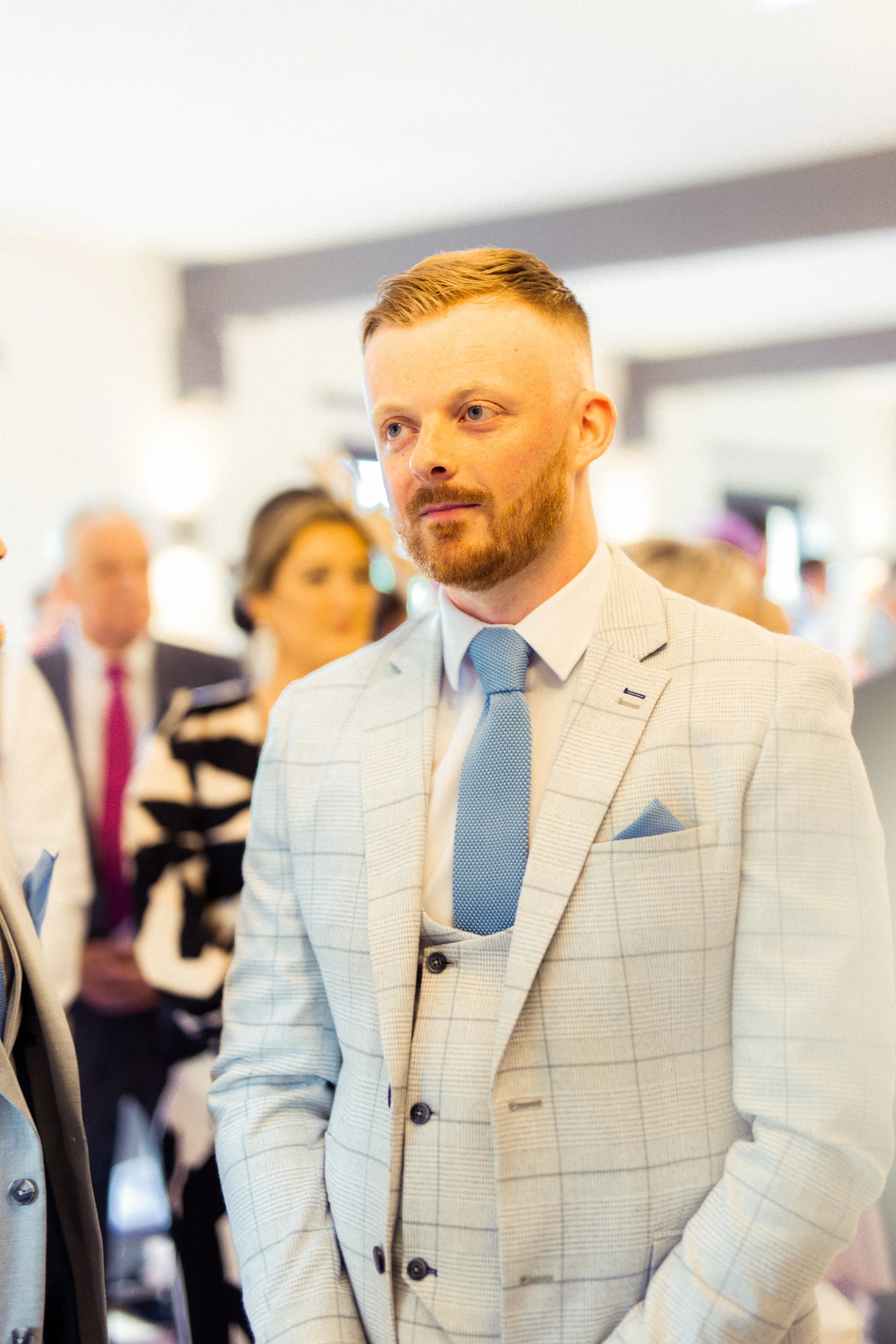A man with a red beard and light brown hair, dressed in a light gray checkered suit, white shirt, and light blue tie, standing in a crowded indoor event, listening attentively.