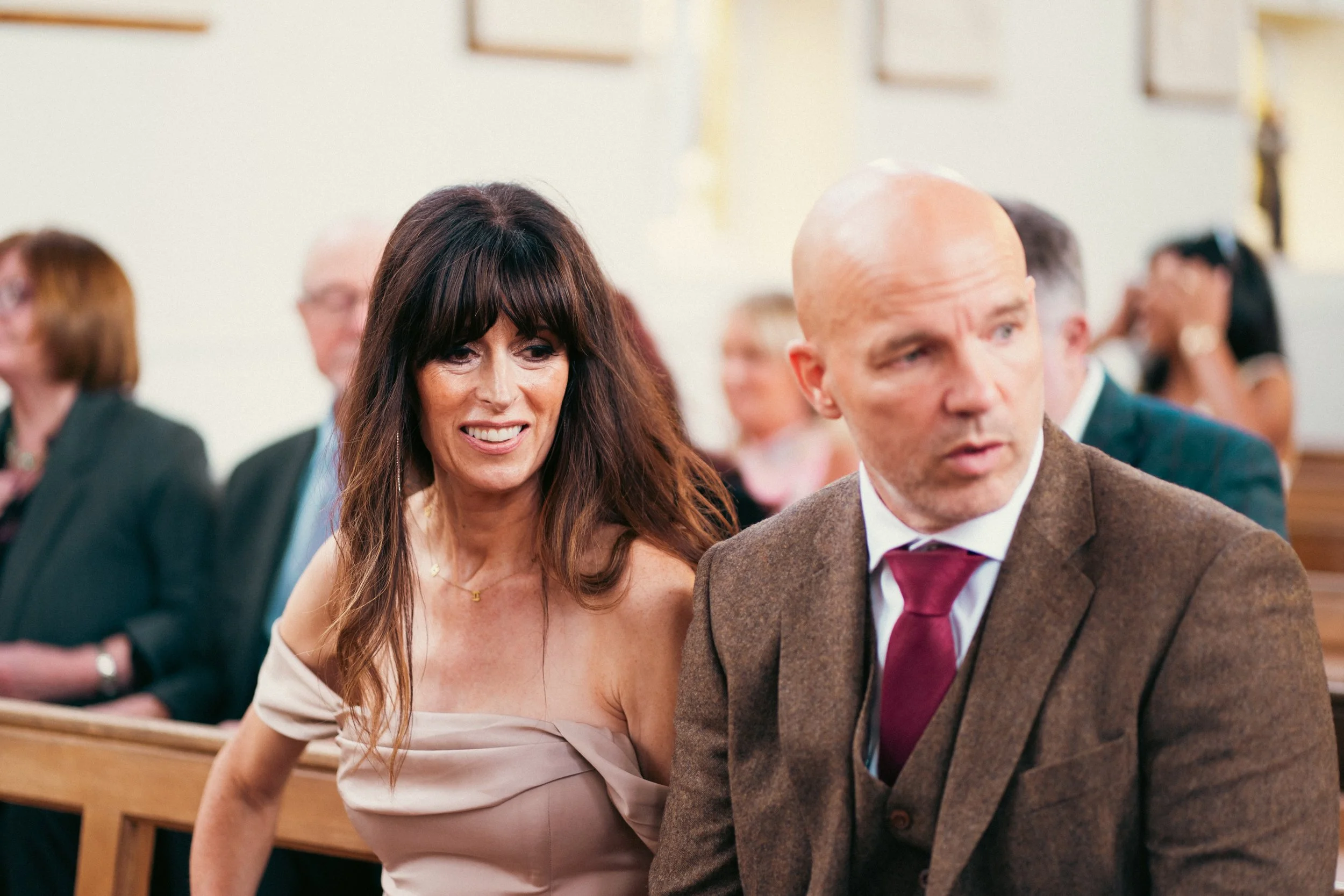A man and woman sitting in a church, dressed formally, with other people seated behind them.
