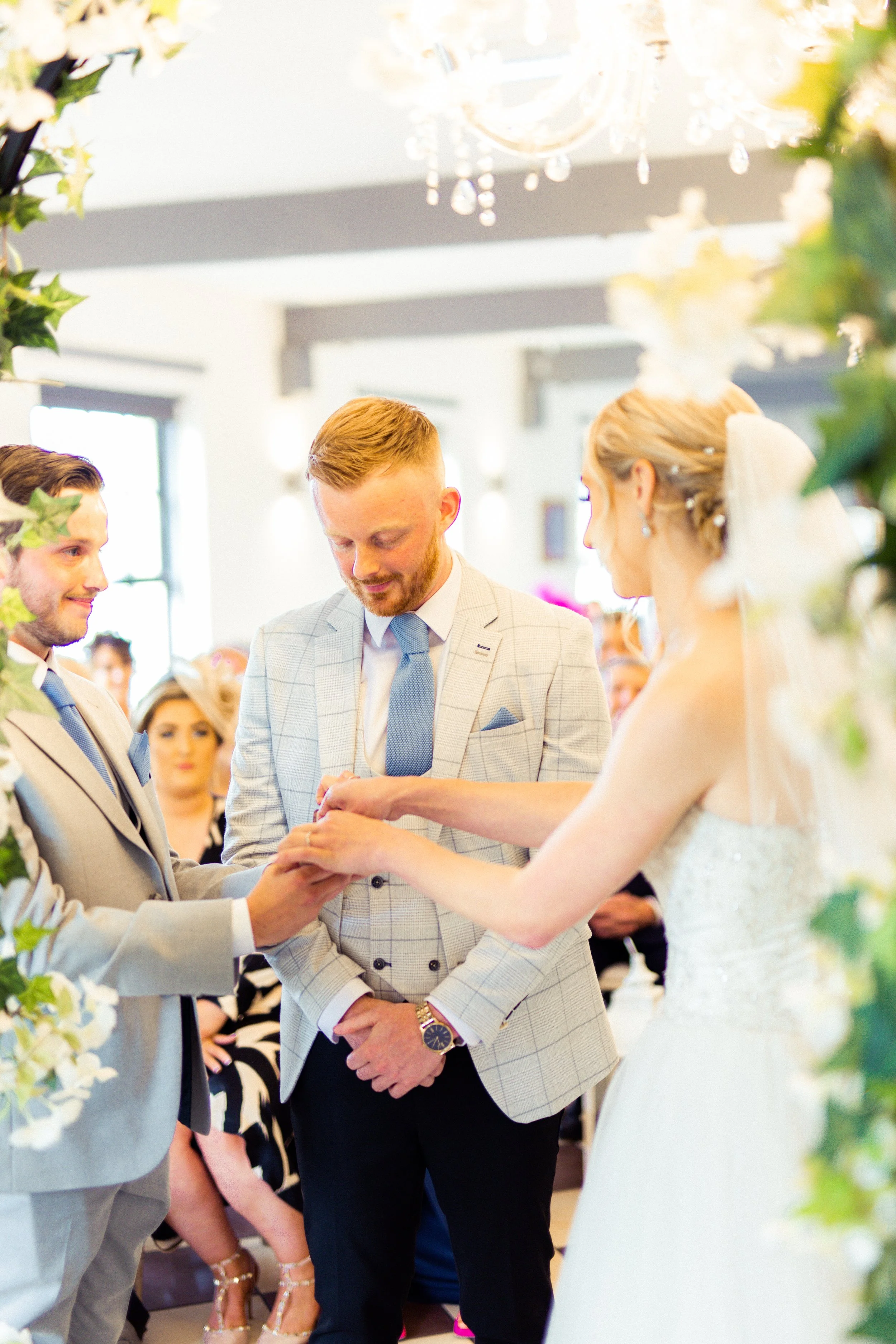 Bride and groom exchanging wedding vows during ceremony, with an officiant helping hold the bride's hand, decorated with greenery and flowers.