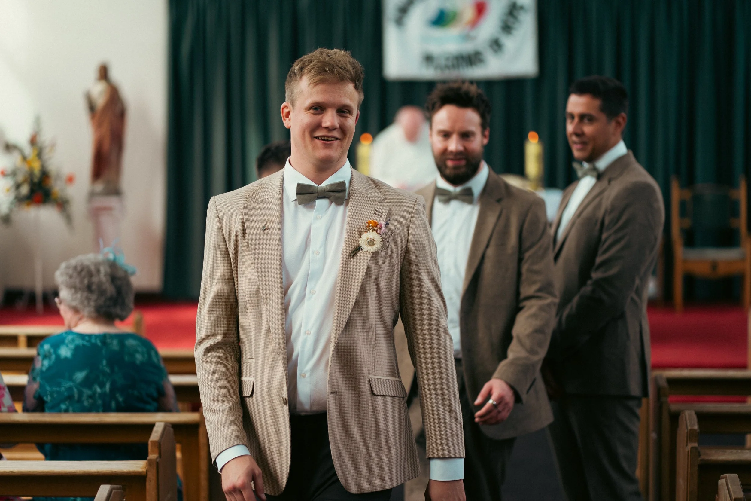 A group of three men in suits and bow ties at a wedding or formal event, standing in front of a stage with a green curtain and a banner, with an older woman sitting in front facing away from the camera.