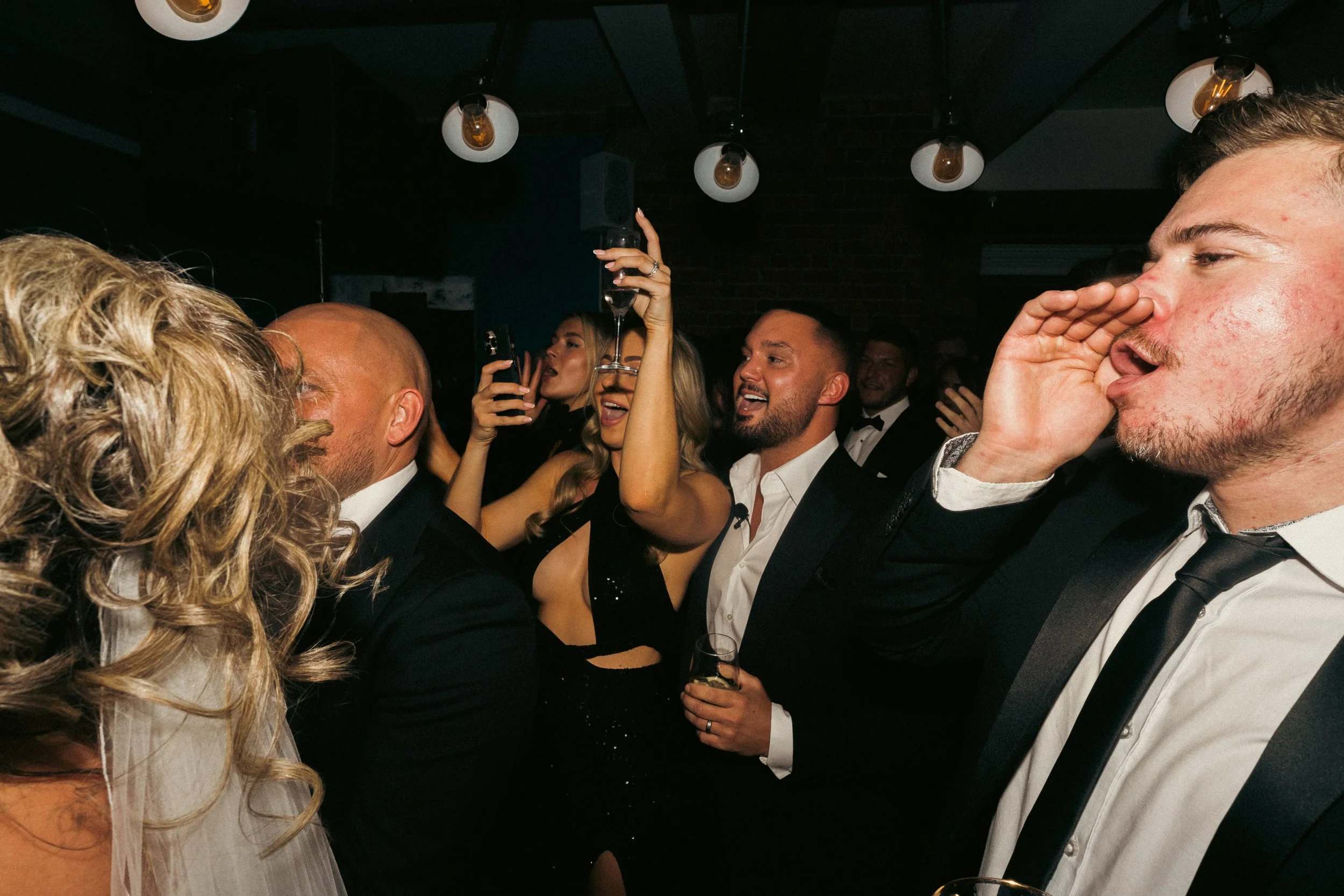 Group of people at a celebration, dressed in formal attire, dancing and enjoying drinks at a party or nightclub.