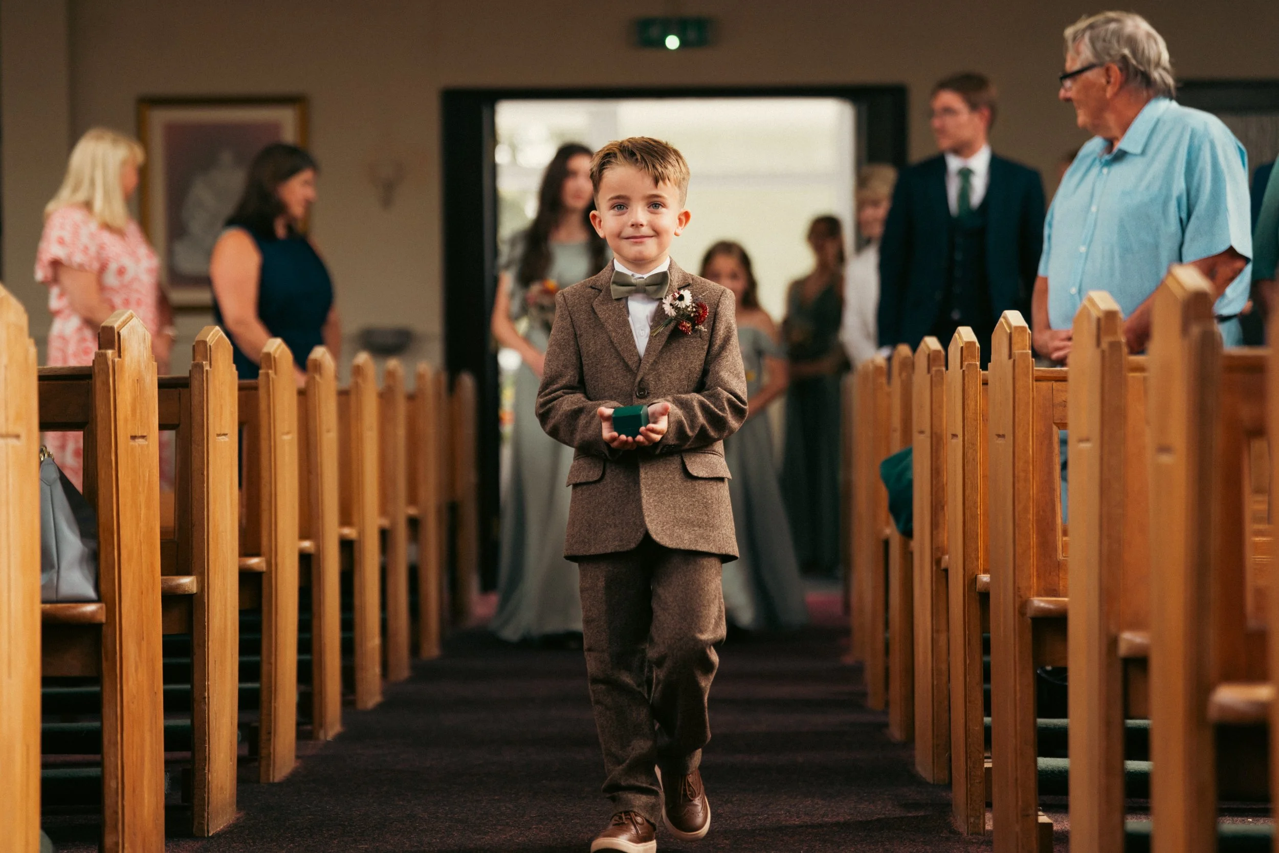 A young boy in a brown suit with a boutonniere walks down the aisle of a wedding ceremony with guests seated on either side.