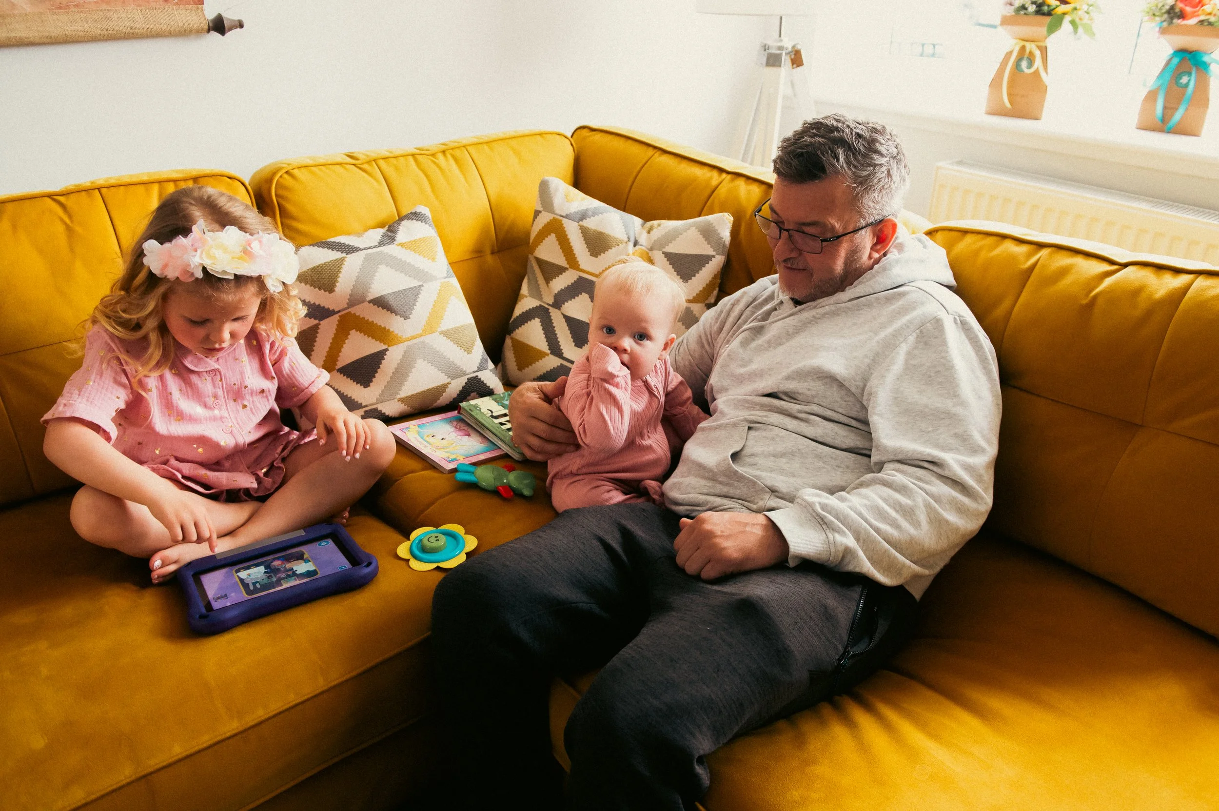 A man sitting on a yellow couch with two young girls and a baby girl. The man is holding the baby girl, who is dressed in pink. The girl on the left is playing with a tablet, and the girl in the middle with a flower crown is watching it.