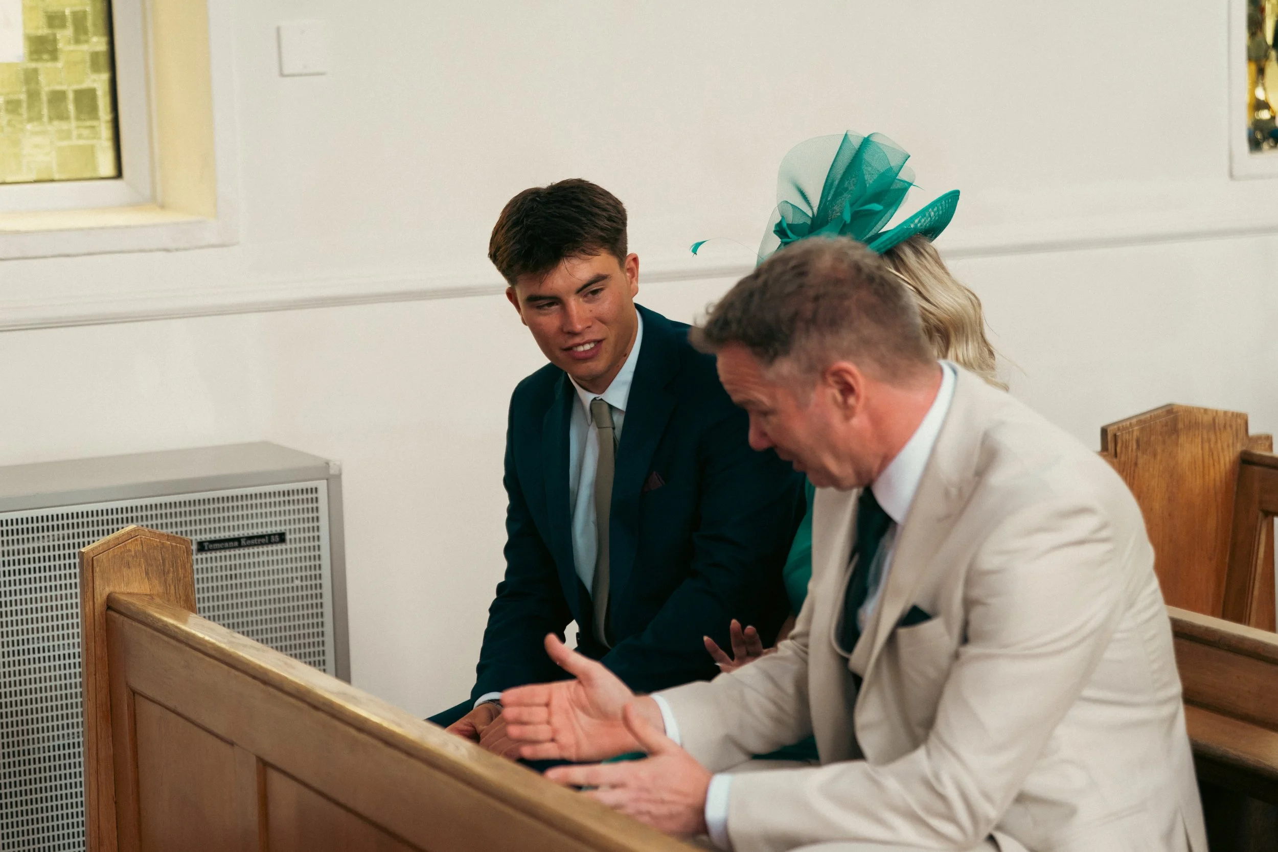 Three people sitting in a church pew engaged in conversation, two men and a woman with teal fascinator, in formal attire.