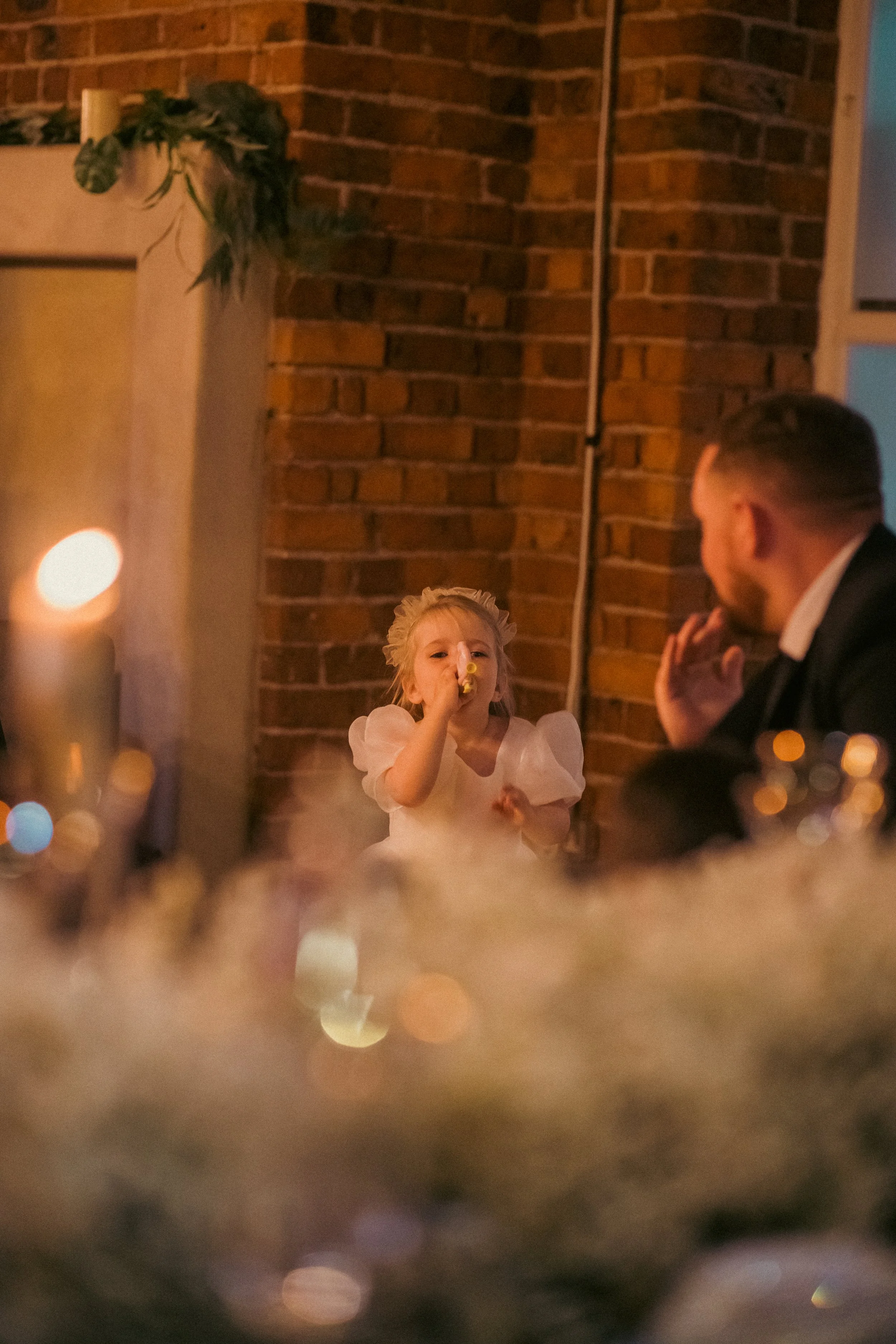 A young girl with blonde hair in a white dress blowing out candles at a birthday celebration, with a man sitting nearby, in a cozy indoor setting with a brick wall background and blurred festive decorations.
