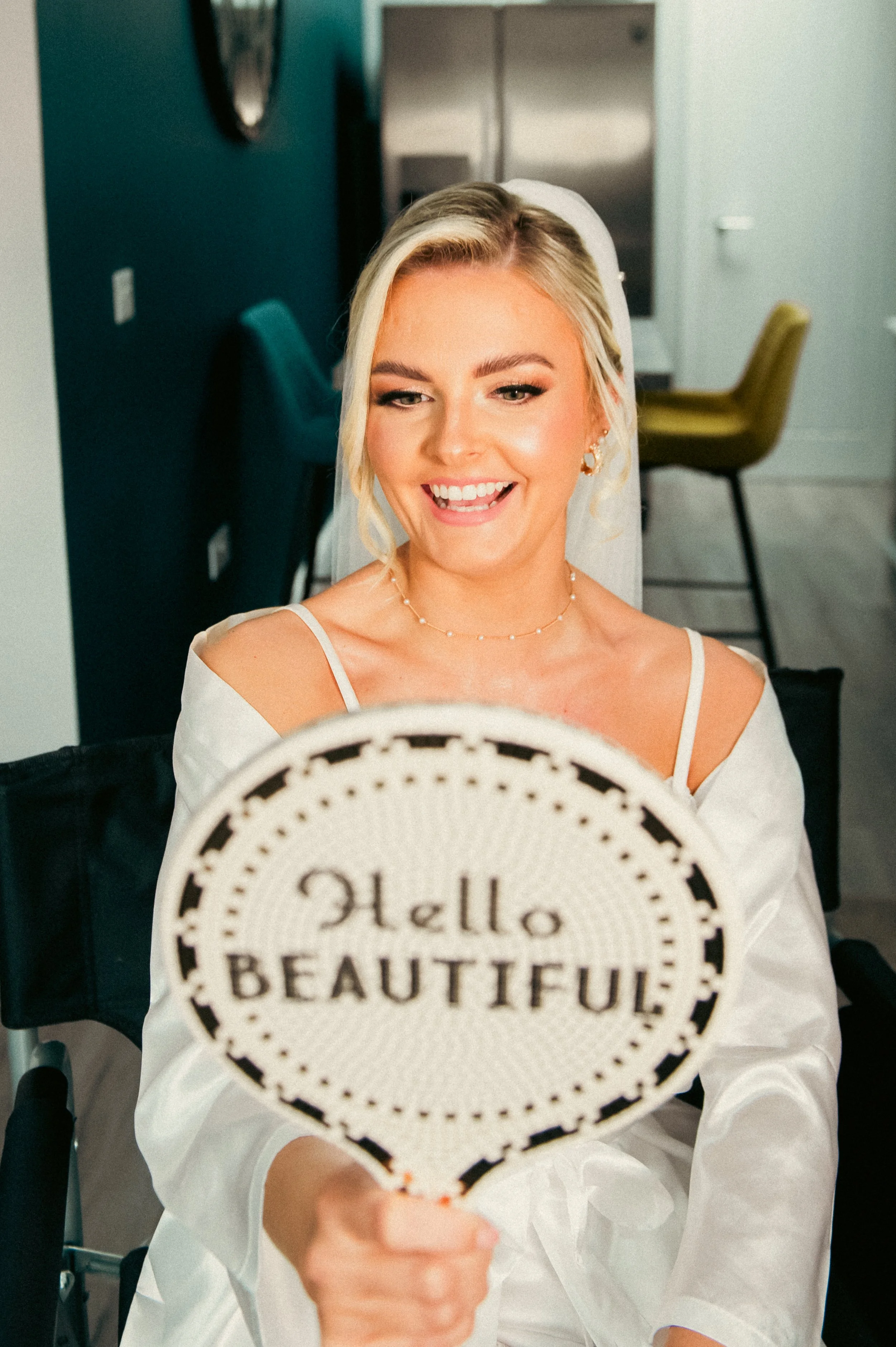 A woman with blonde hair and jewelry holding a sign that says 'Hello Beautiful' while smiling.