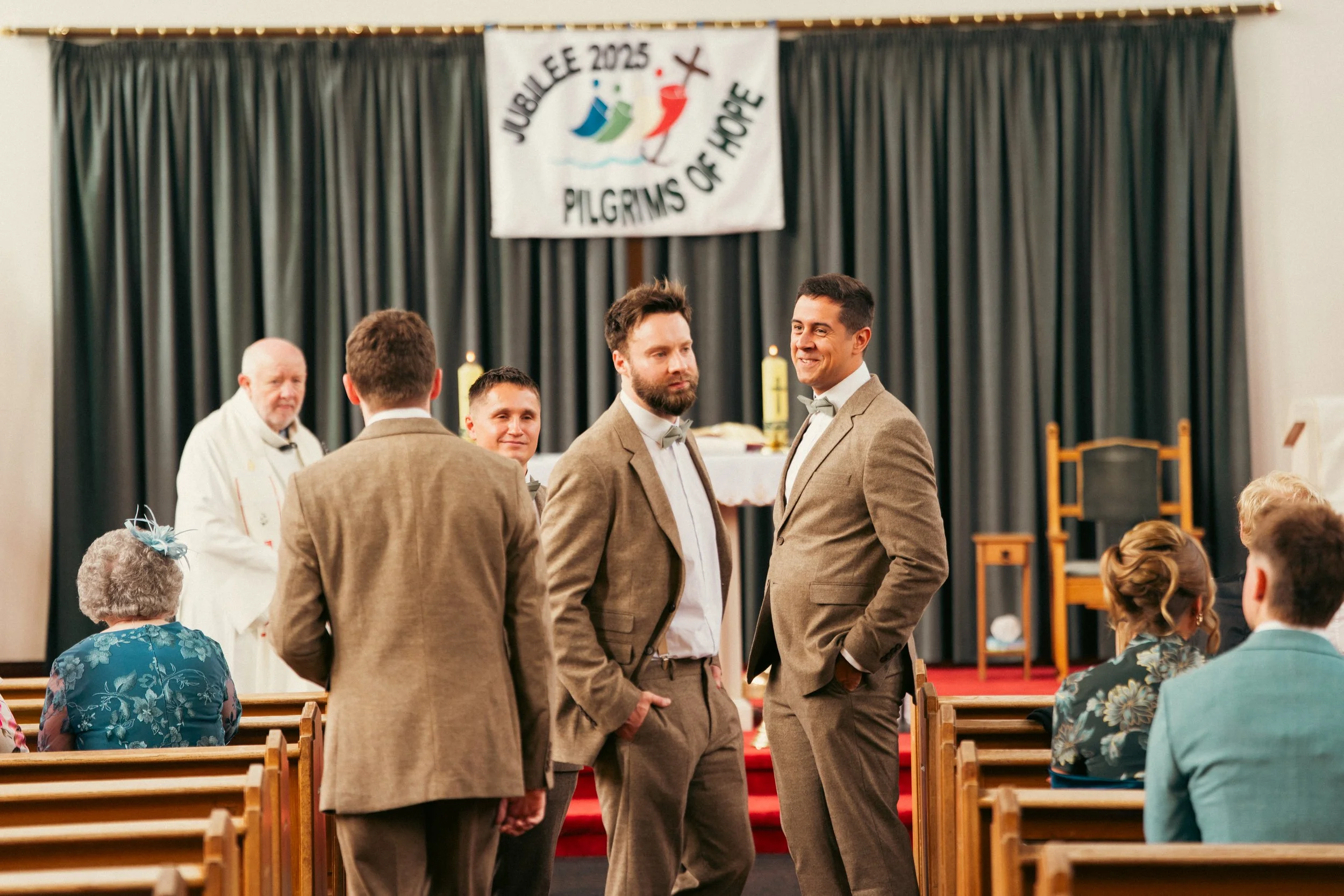 Wedding ceremony in a church with five men in suits, two women in floral dresses seated, and a priest in the background. A banner reads "Jubilee 2025 Pilgrims of Hope."