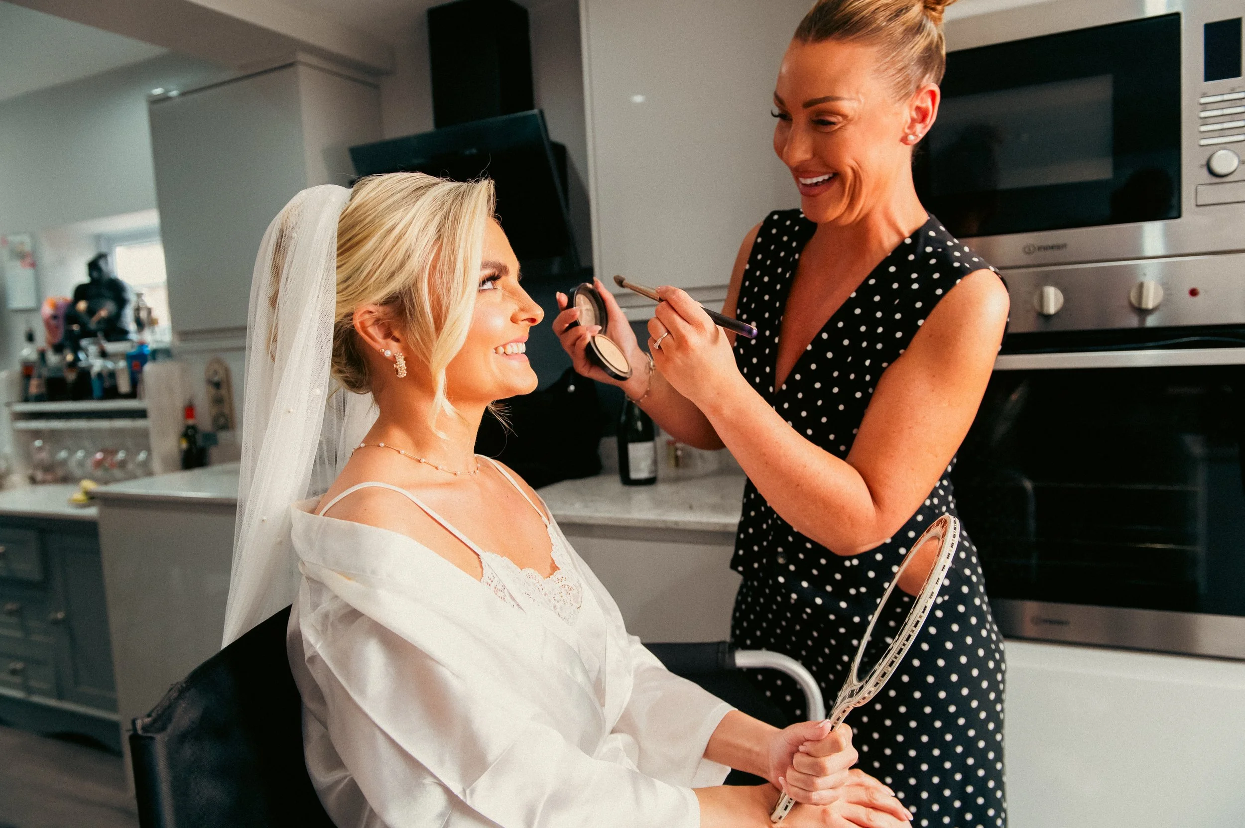 A bride getting makeup done in a kitchen by a woman holding a makeup brush and a mirror.