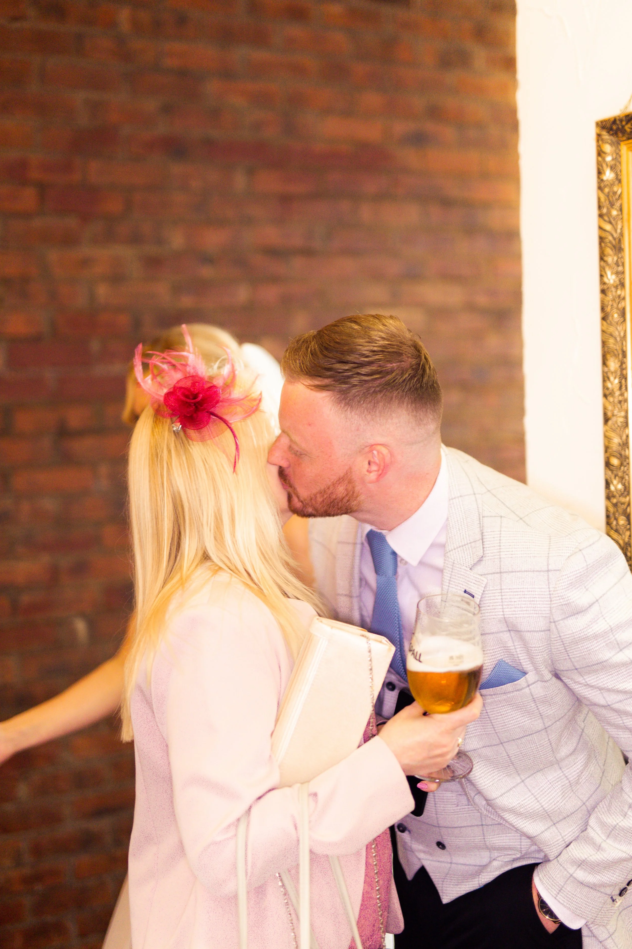 A man and woman kissing at a social event, the woman holding a glass of beer and a notebook, with a brick wall in the background.