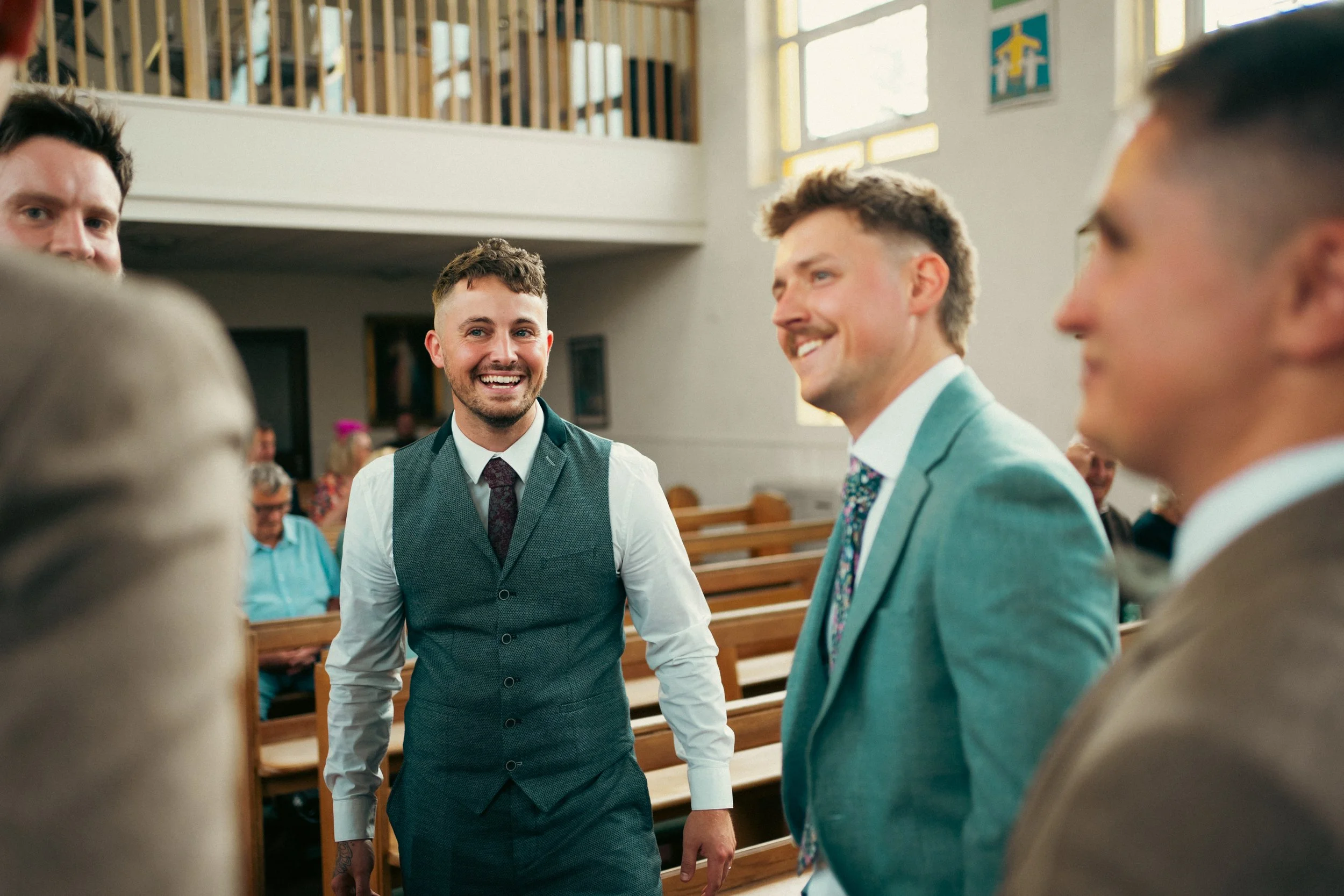 Group of men dressed in suits and vests smiling and talking inside a church during a wedding.