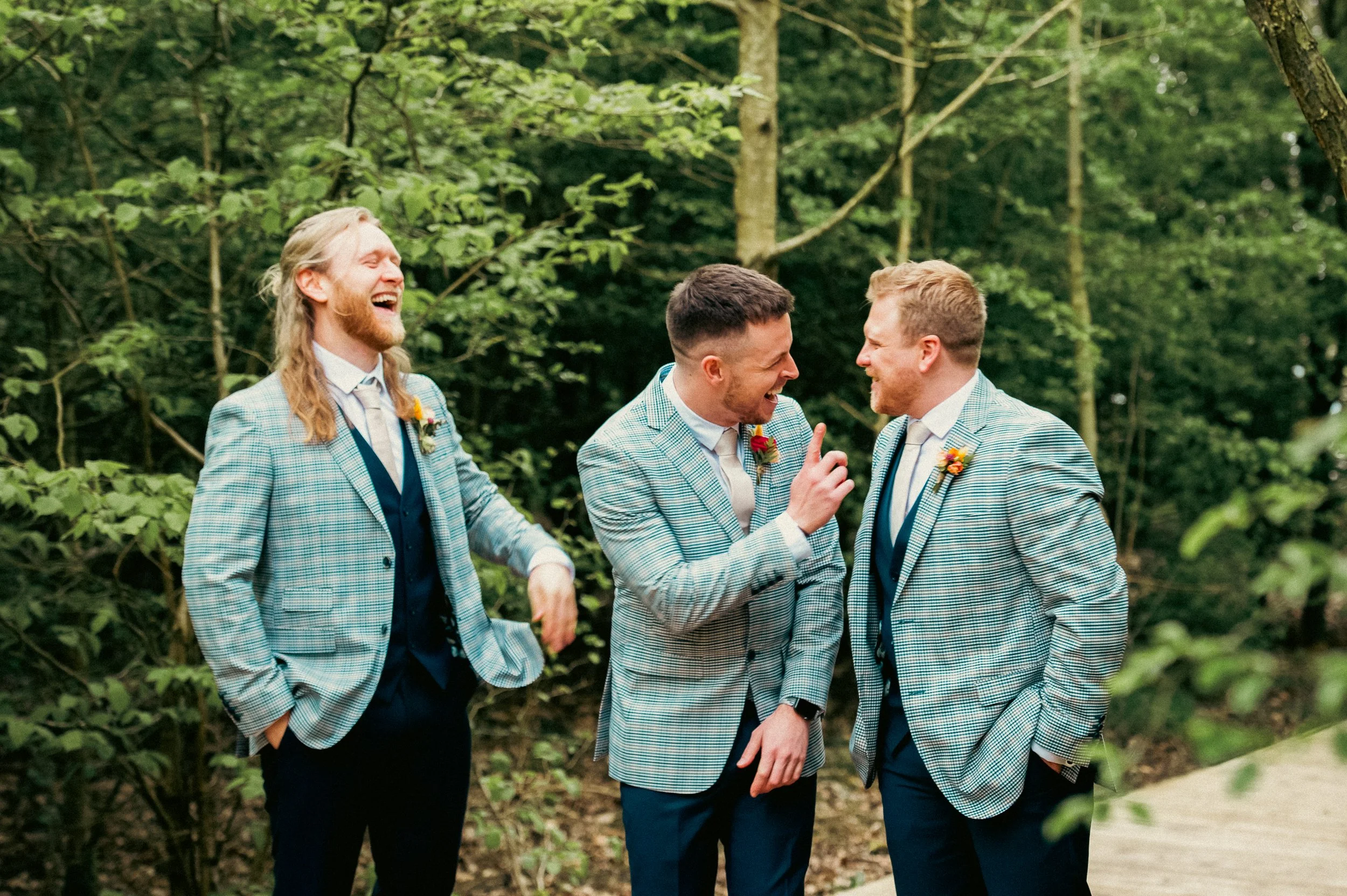 Three men in matching plaid suits and boutonnieres laughing and talking in a wooded outdoor setting.