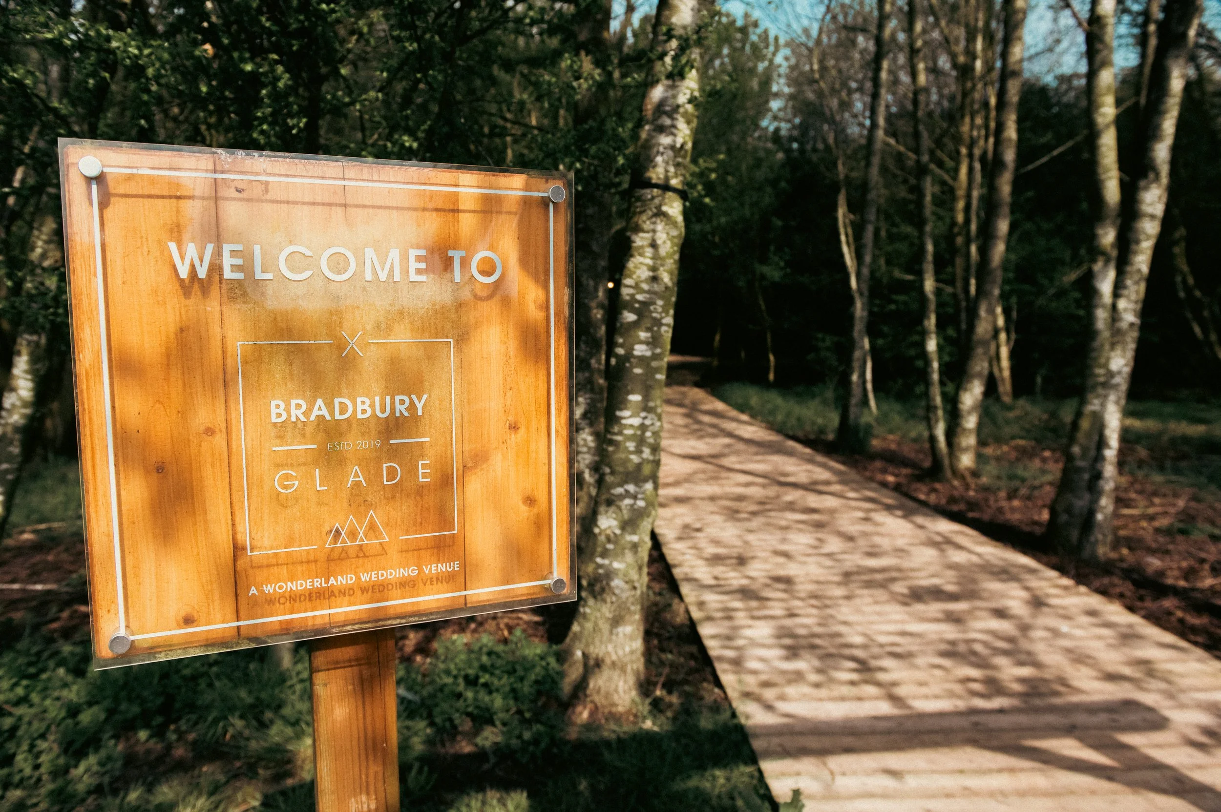 Wooden welcome sign for Bradbury Glade, a wedding venue established in 2019, with a pathway surrounded by trees leading into a wooded area.