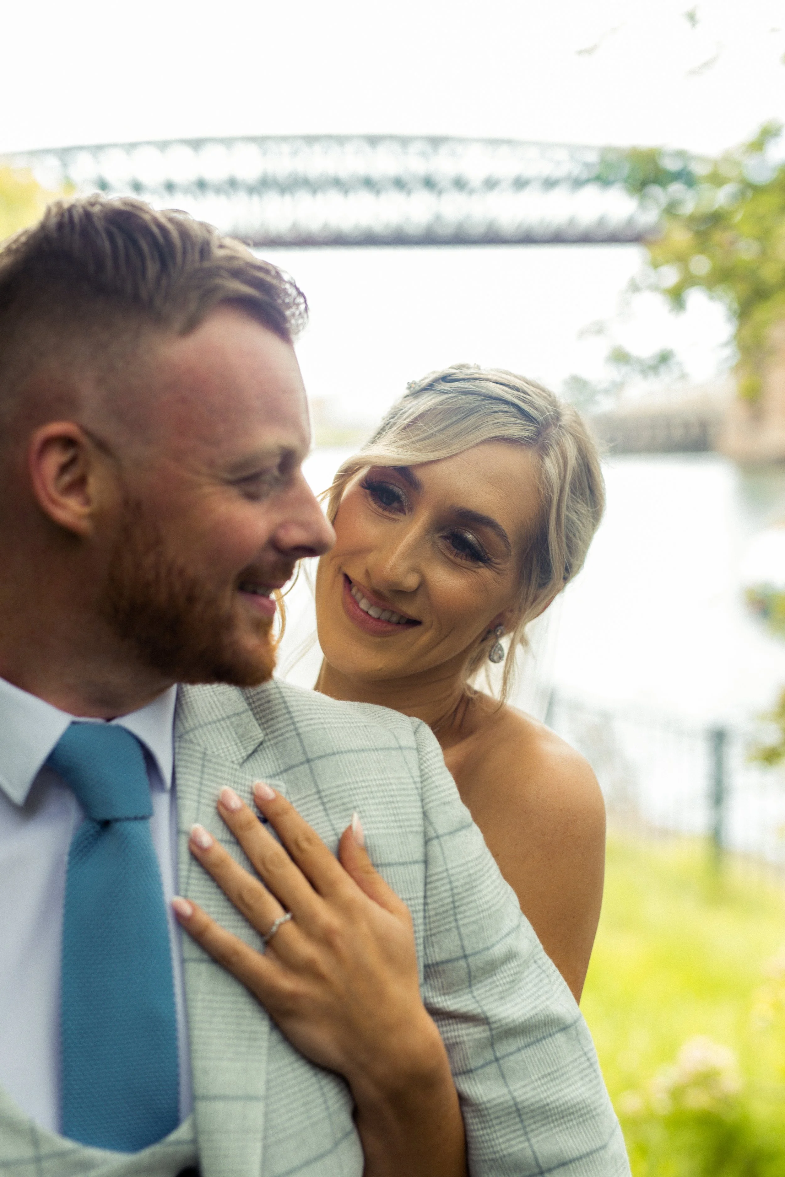 Close-up of a smiling bride looking at a groom near the river with a bridge in the background