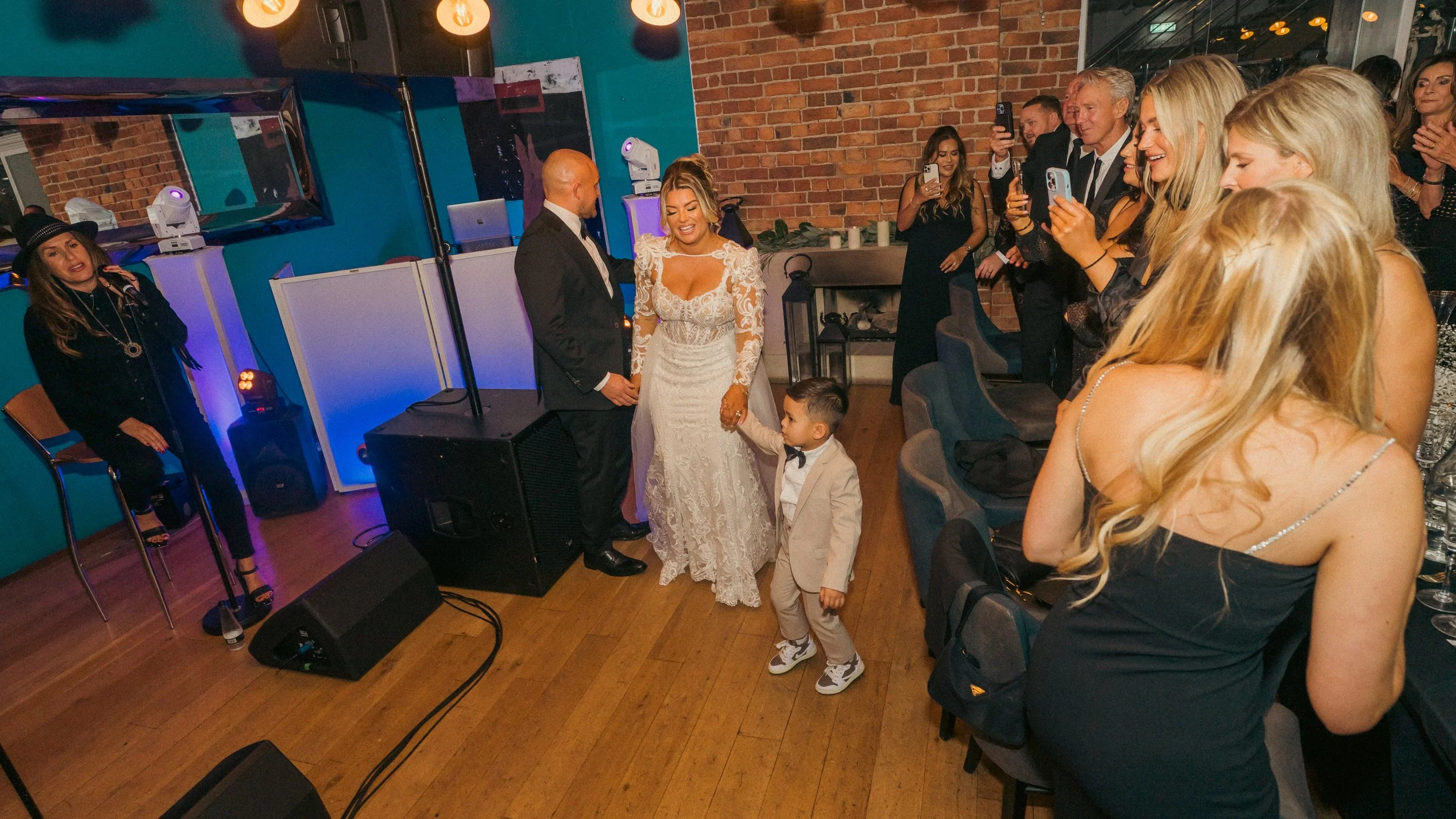 Wedding reception with bride, groom, and young boy dancing in front of seated guests who are taking photos and videos.
