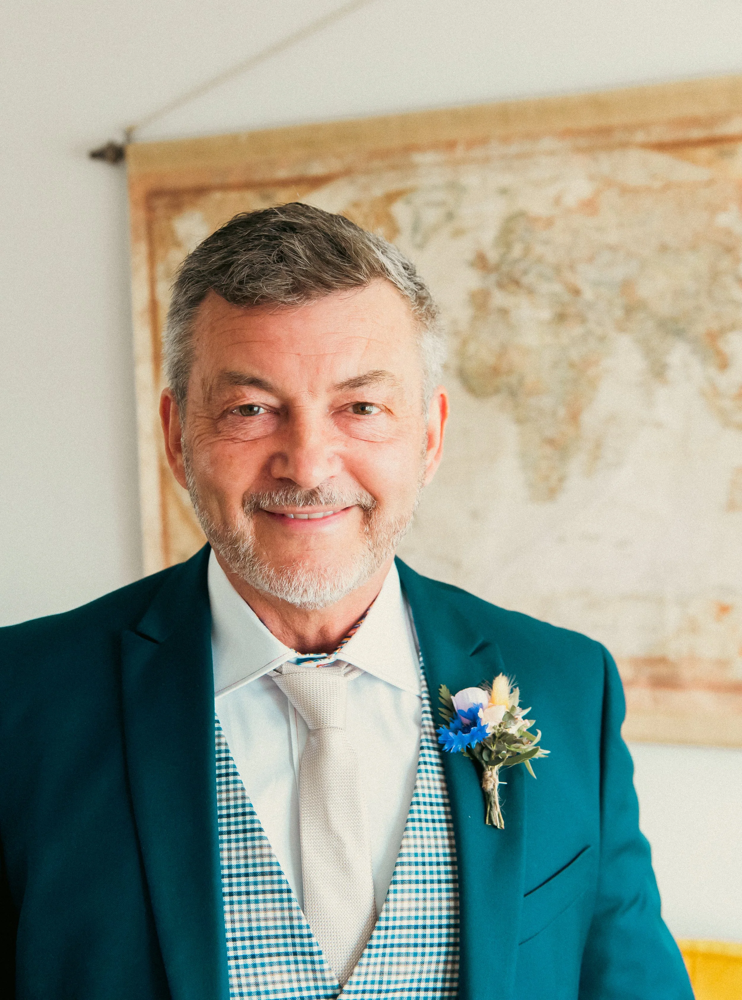 A smiling older man dressed formally in a tuxedo with a white tie, a checkered vest, and a boutonniere with blue and white flowers, standing in front of a world map.