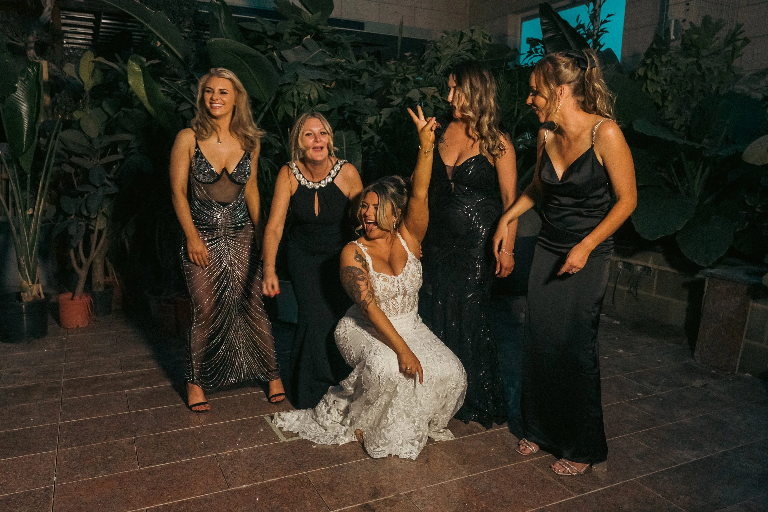 Group of six women dressed in formal evening gowns, posing together indoors with large plants in the background. One woman in a white lace dress is kneeling in front, making a peace sign with her hand. The other women are standing around her, smiling