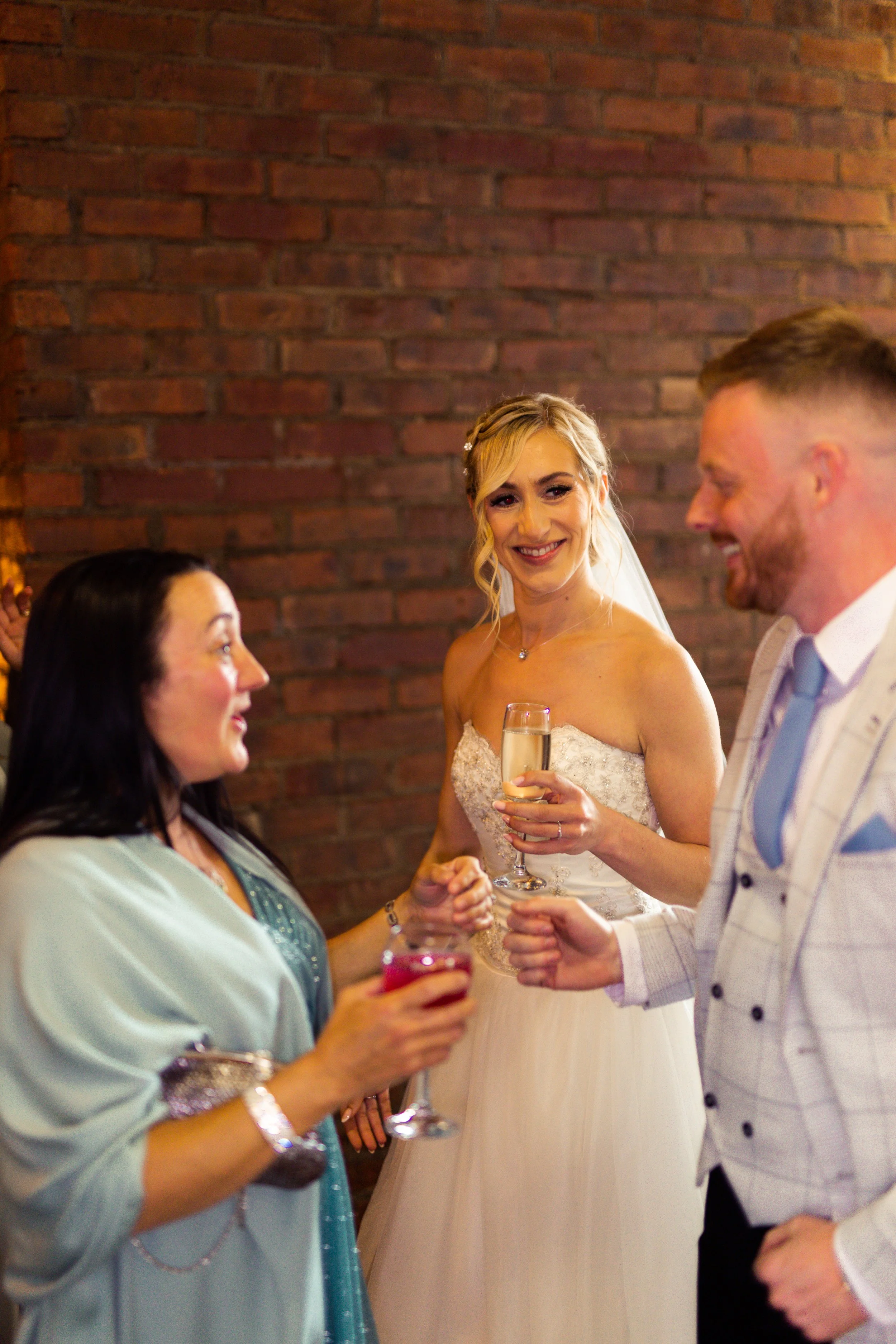 A bride with blonde hair in a strapless wedding gown holding a champagne glass, smiling at a woman with dark hair in a green dress, who is holding a red drink, while a man in a light-colored suit jacket and tie looks on and smiles, against a brick wa