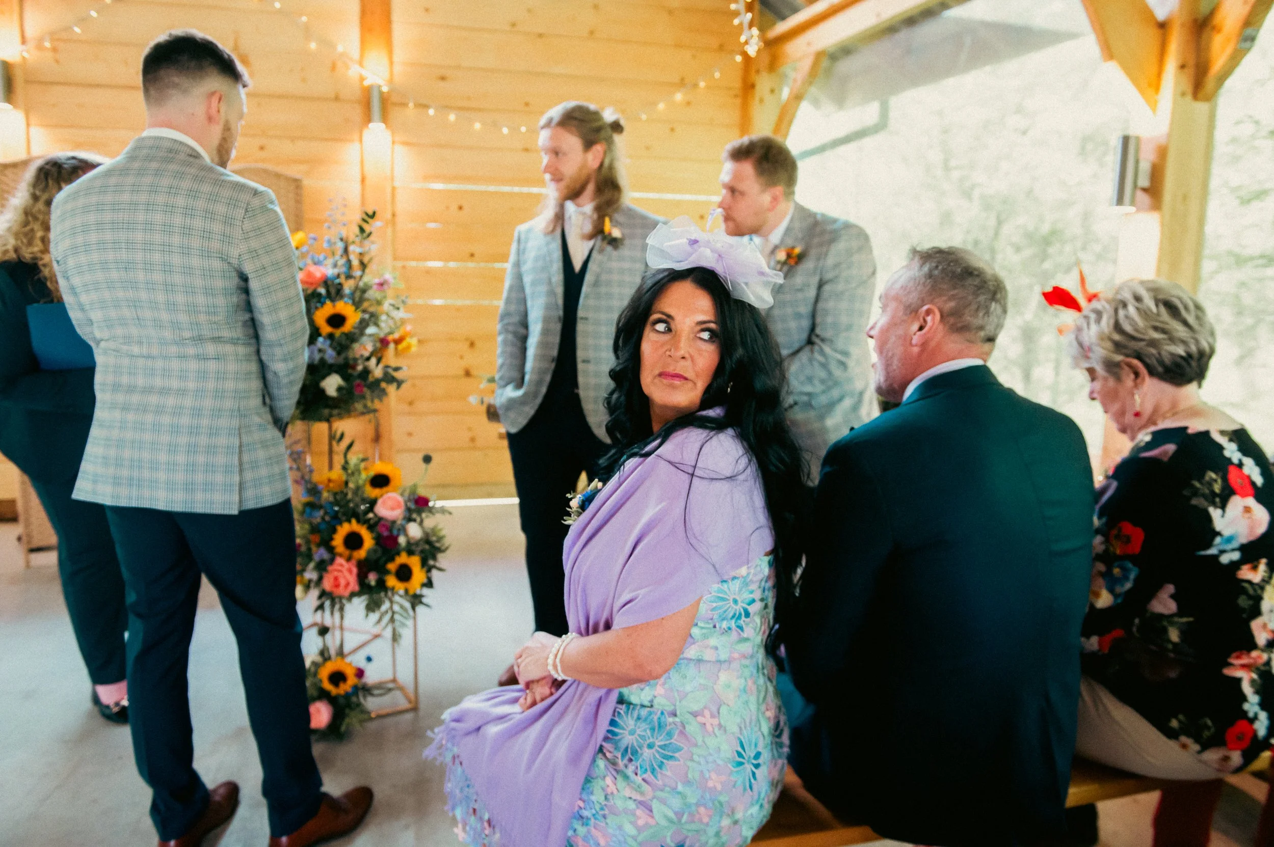 People seated in a wooden chapel decorated with flowers, attending a wedding ceremony. The woman in the foreground is wearing a lavender dress and fascinator, looking to the side. Other guests are engaged in conversation or listening to the ceremony.