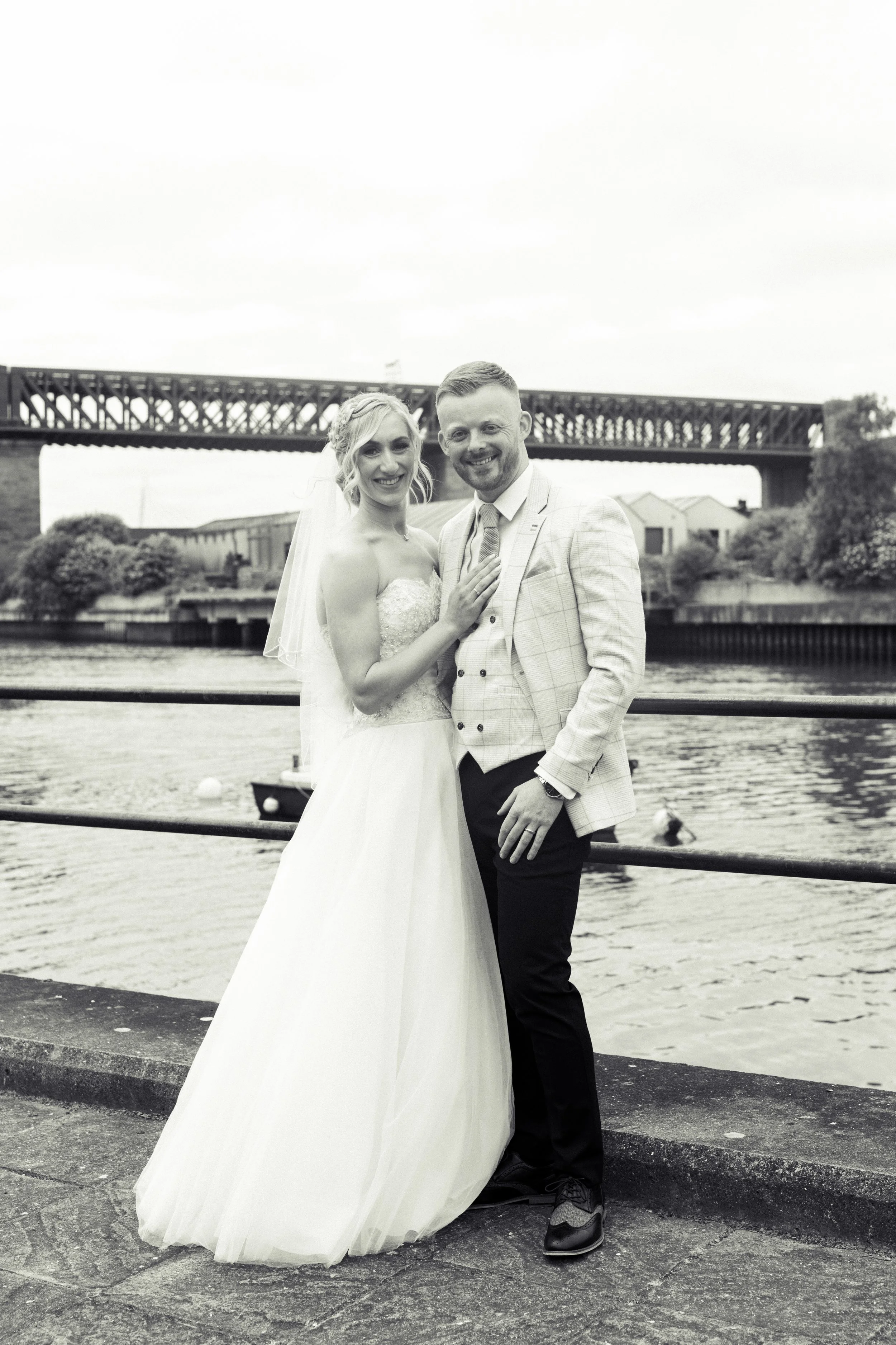 A black and white photo of a bride and groom standing near a river with a bridge in the background, smiling at the camera.