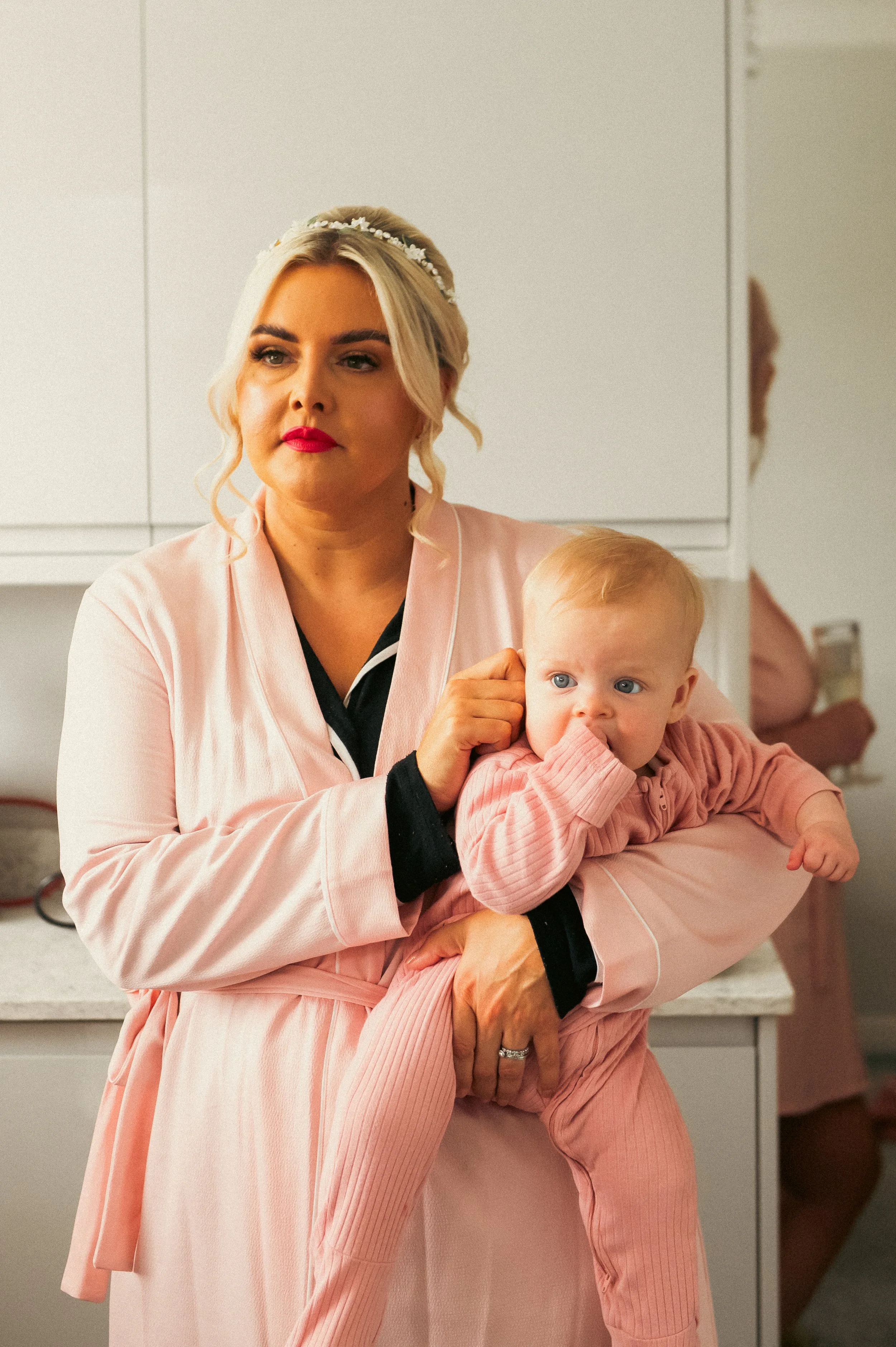 A woman with blonde hair, wearing a pink robe with black pajamas underneath, holding a young child dressed in pink pajamas. They are standing in a kitchen with white cabinets.