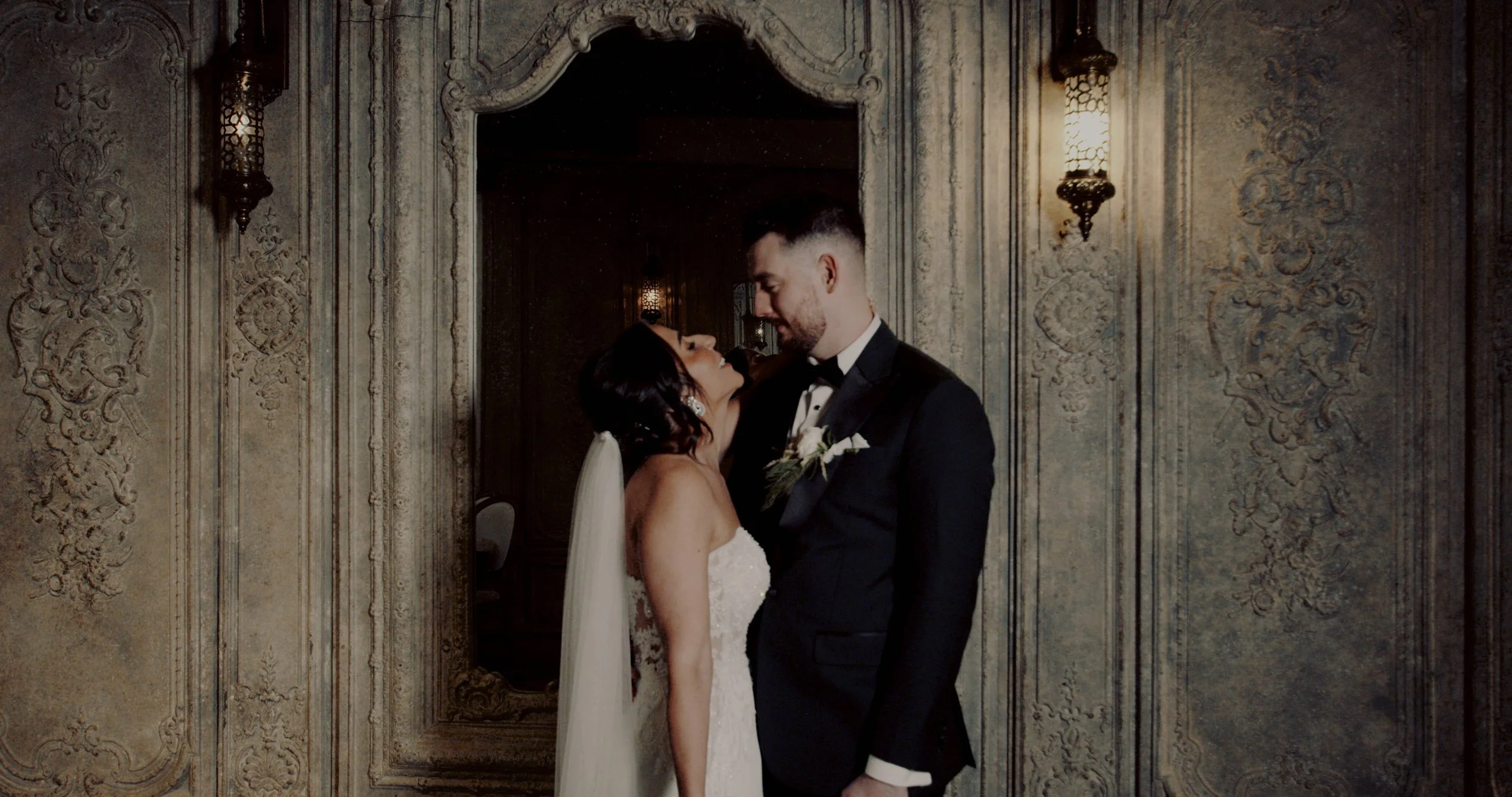 A bride and groom in wedding attire standing close and looking into each other's eyes in front of an ornate, vintage wall with a mirror and wall sconces.