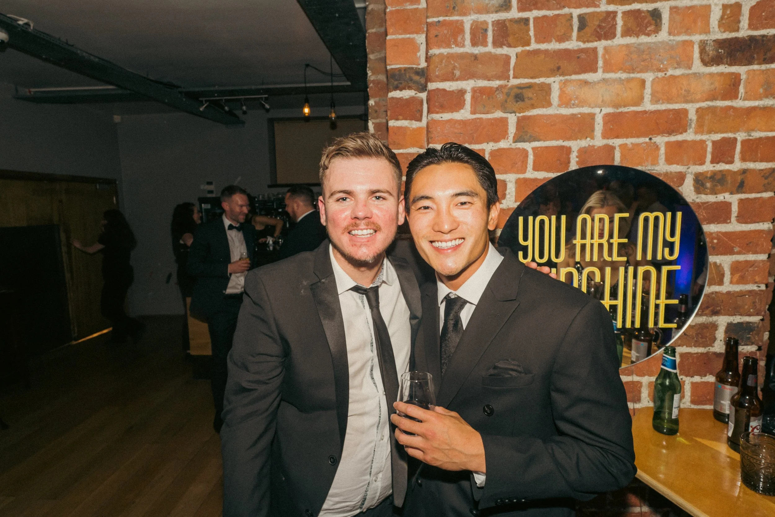 Two men in suits smiling for a photo at a party, with a bar and other guests in the background, and a sign that reads 'You are my sunshine' on a brick wall.