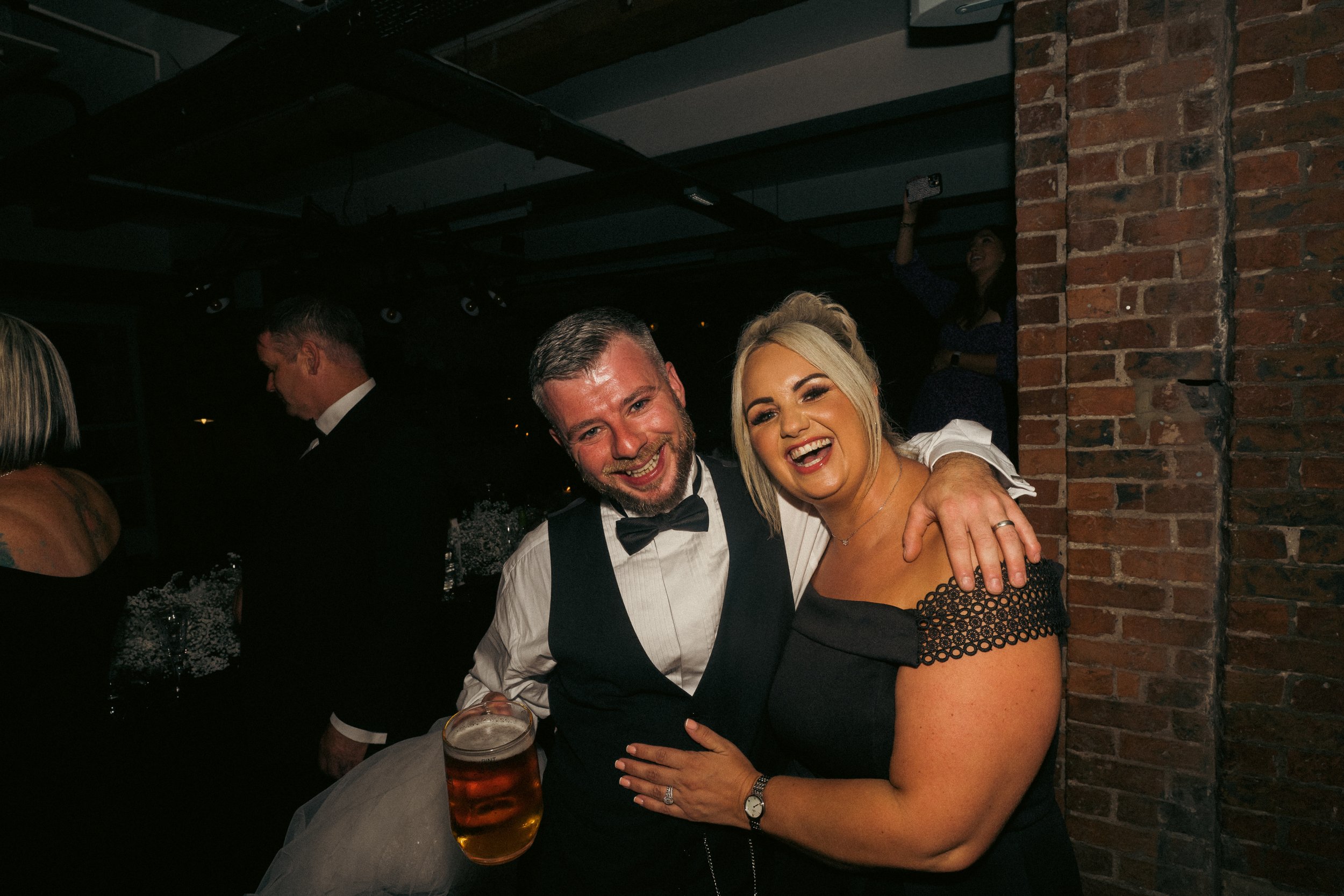 A man and woman smiling and celebrating at a party, with the man holding a glass of beer, standing close together, with a brick wall in the background.