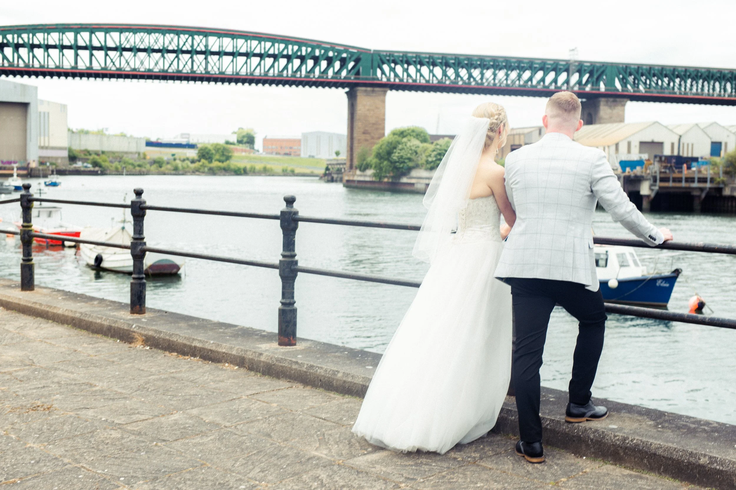 A bride and groom stand by a waterfront railing, overlooking boats in the water with a bridge and industrial buildings in the background.