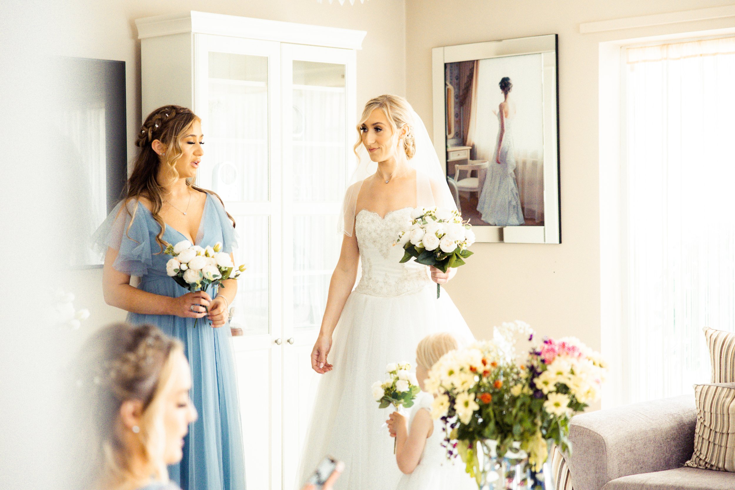 A bride in a wedding dress holding a bouquet, standing in a room with two young girls, one in a blue dress and one with blonde hair, holding flowers. The room has a mirror, a cabinet, and a framed picture of a woman in a gown.
