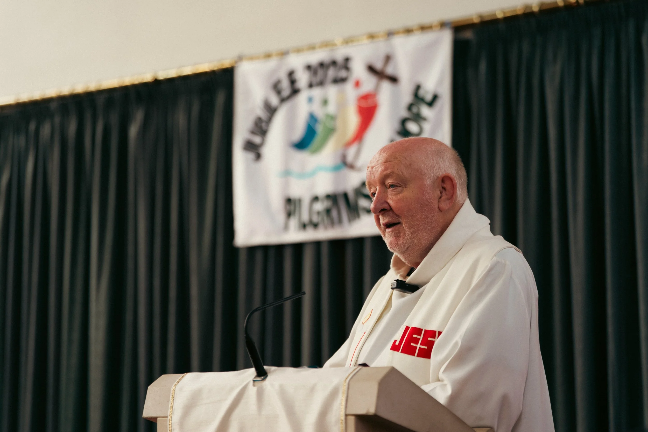 A man with a white beard wearing a white robe with red letters speaking at a podium with a black microphone. Behind him is a banner with colorful water-themed figures and text.