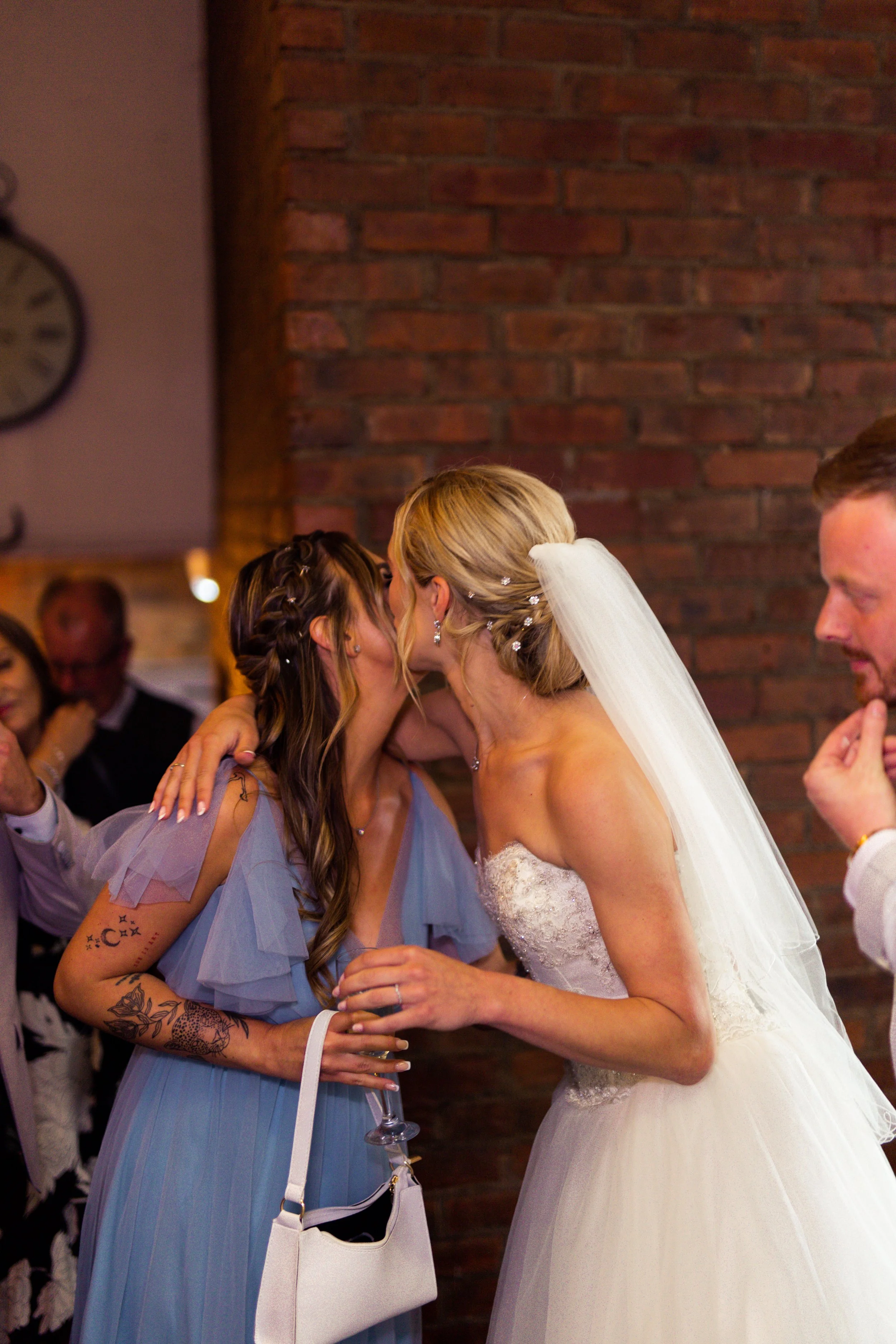 A bride and a woman share a kiss at a wedding reception, with a stone wall in the background and guests watching.